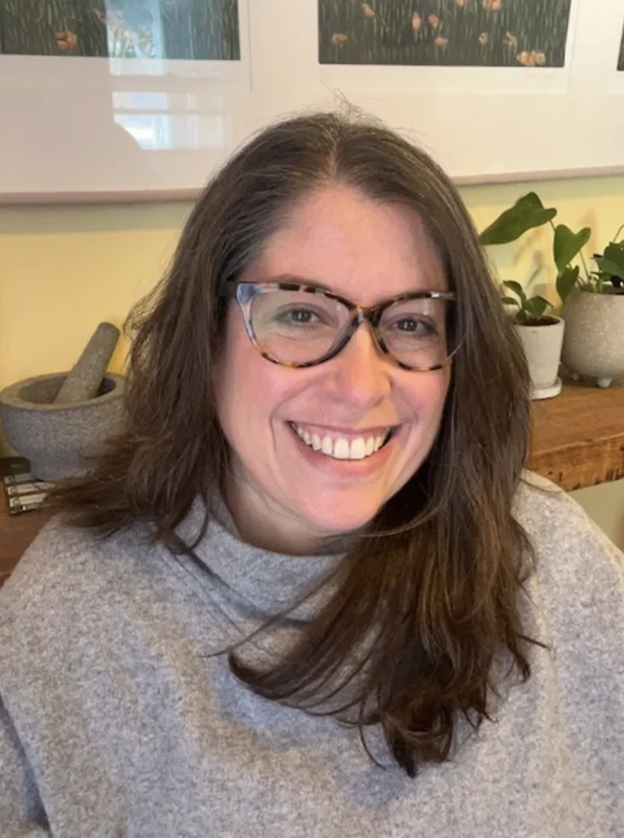 Smiling woman wearing glasses and a gray turtleneck sweater, sitting in a cozy indoor space with plants and framed pictures in the background.