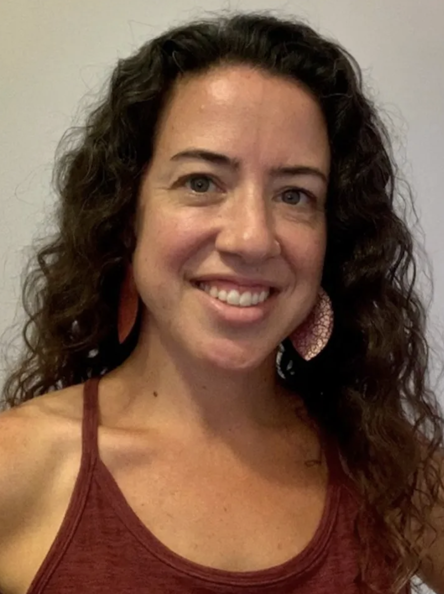 A smiling woman with curly dark hair, wearing a maroon tank top and large earrings, stands against a plain light-colored wall.