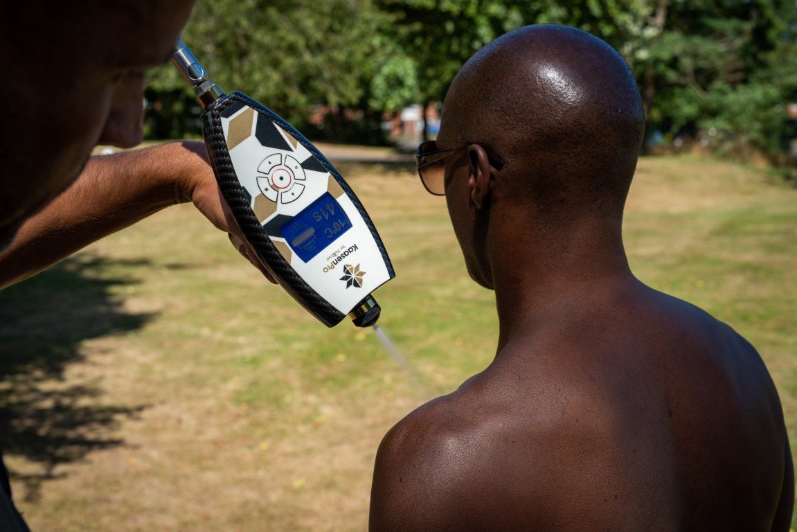 A person holding a portable breathalyzer device in front of a man with dark skin and sunglasses, outdoors on a sunny day, with trees and grass in the background.