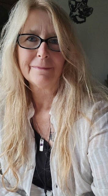 A woman with long blonde hair and glasses taking a selfie indoors, with a dog illustration on the wall behind her.