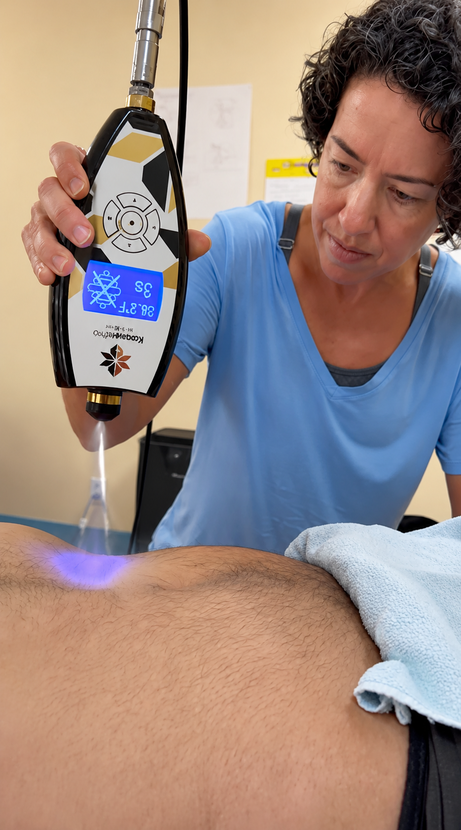 A healthcare professional using a handheld device to perform laser therapy on a patient's back. The device displays the text '3.5' and '34.8'. The patient is lying on their stomach with a towel over their lower back.