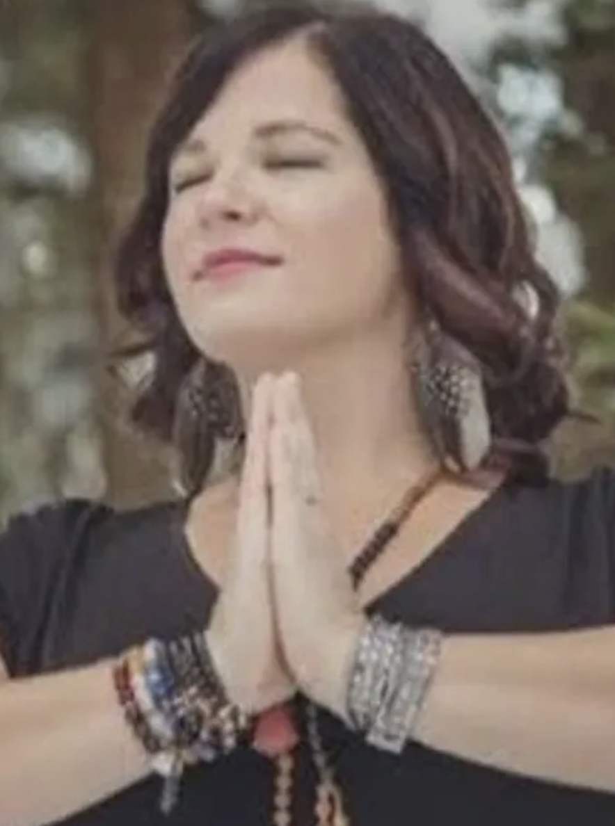 Woman with dark brown hair in loose waves, smiling with eyes closed, hands pressed together in prayer or greeting, wearing large earrings, multiple bracelets, and a black top, outdoors with blurred greenery background.