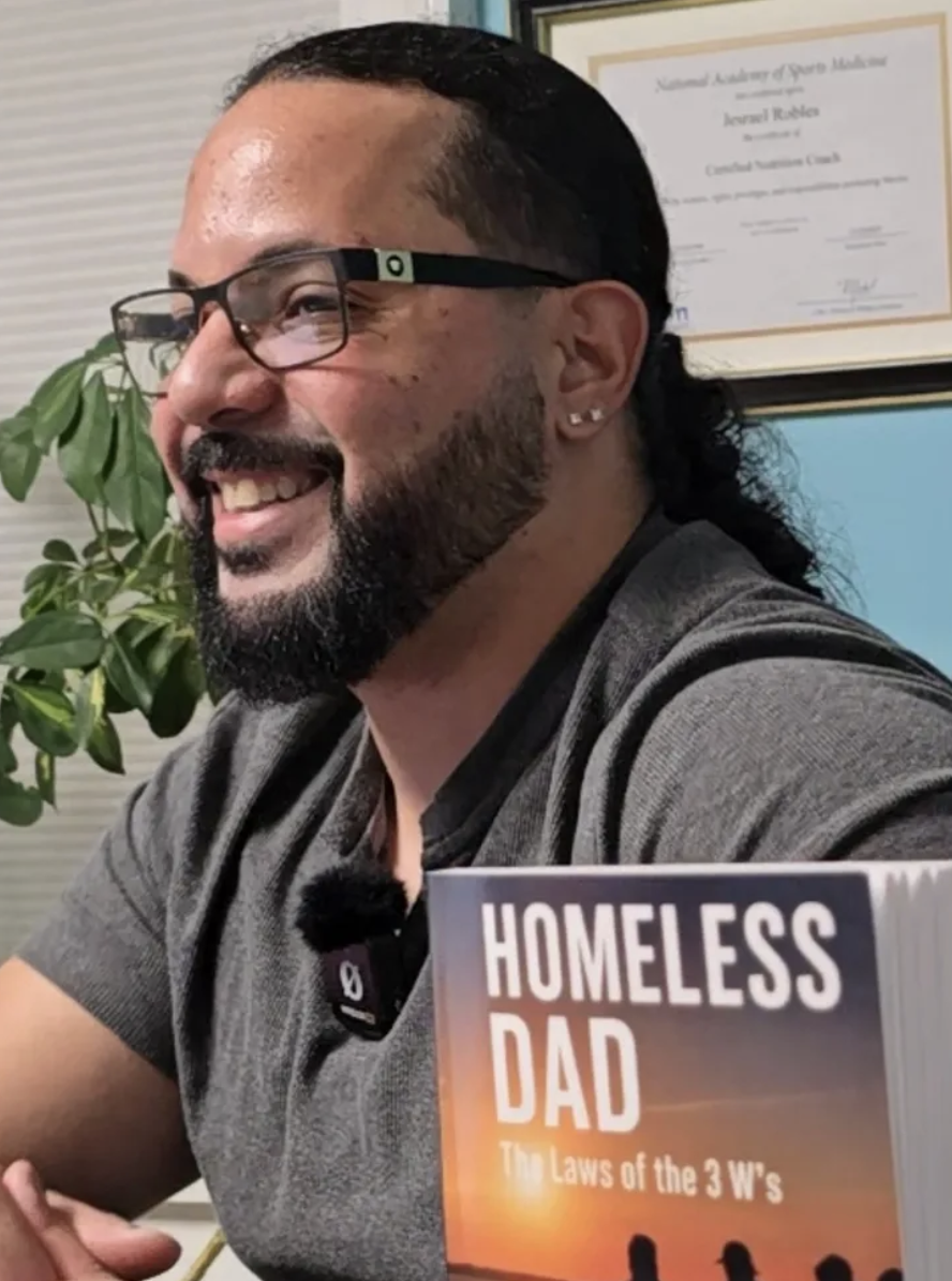 A man with glasses, a beard, and earrings, sitting indoors. He is smiling, and on the table in front of him is a book titled "Homeless Dad," with the subtitle "The Laws of the 3 W's". There is a framed certificate or diploma hanging on the wall behind him and a green potted plant nearby.