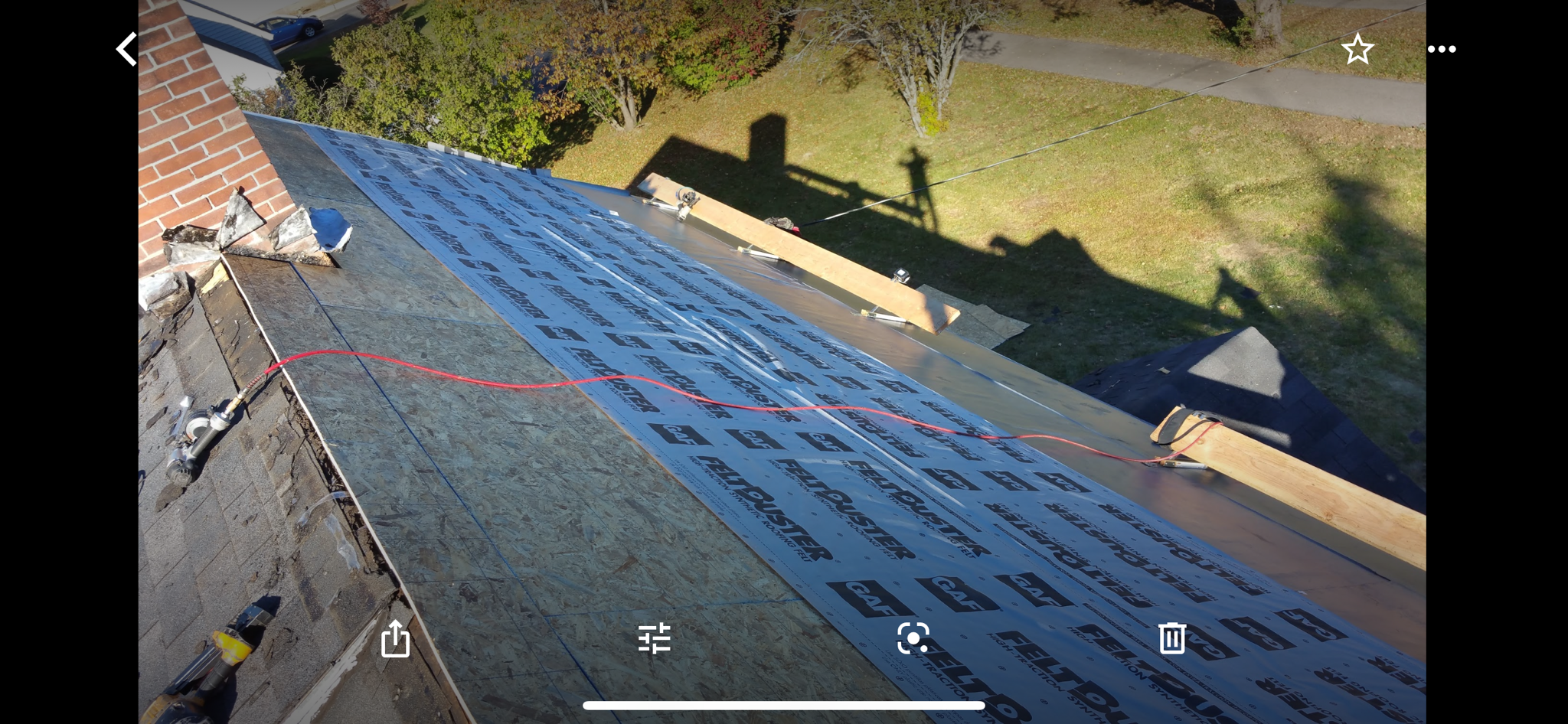 Rooftop undergoing renovation with installation of roofing underlayment, over grey shingles. Tree-lined street visible in background.