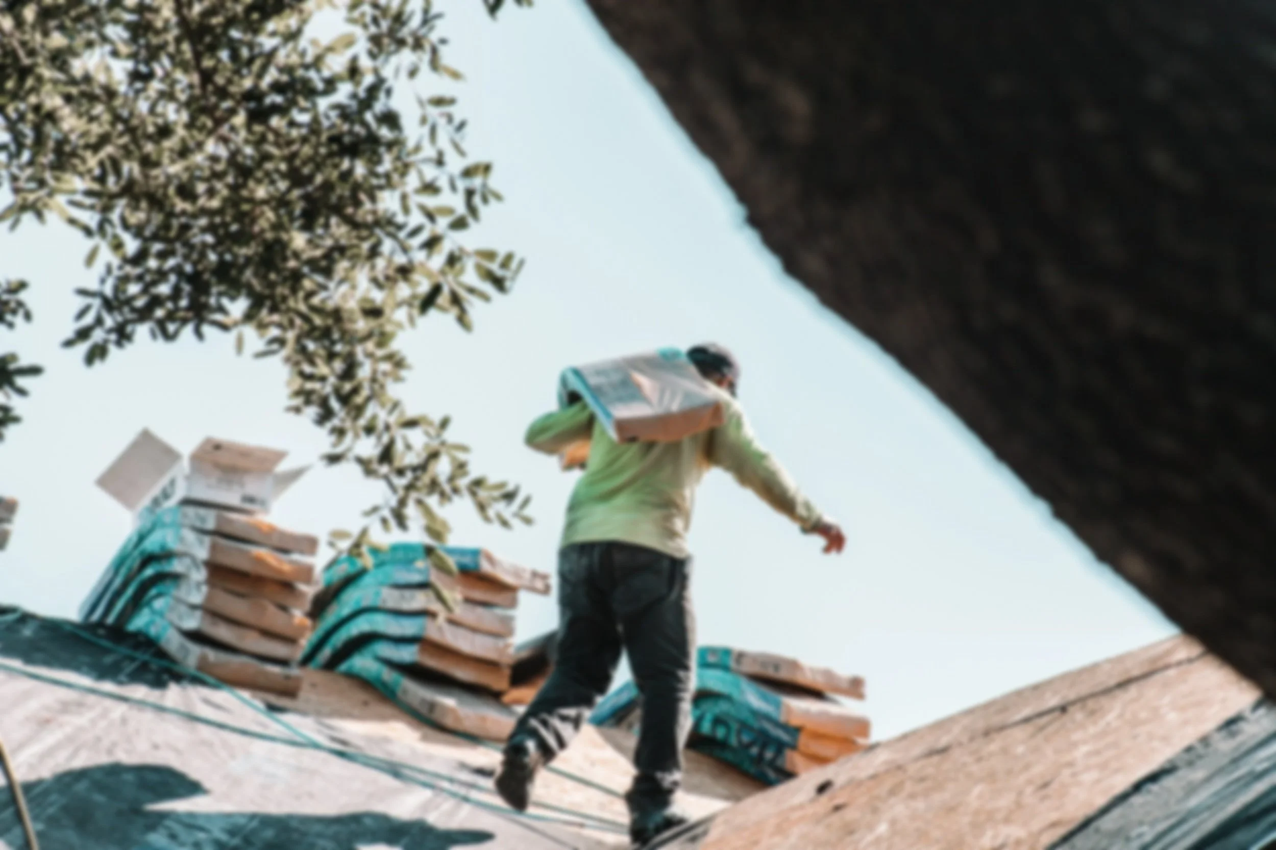 Professional roofing contractor installing asphalt shingles on a residential roof with stacked shingles and trees in the background.