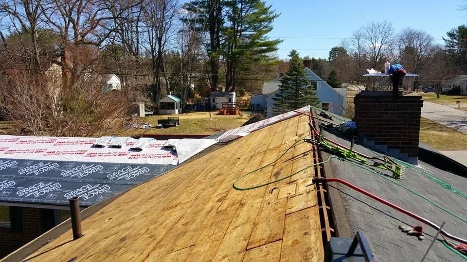 View from a roof under construction, showing a newly installed wooden section and a brick chimney. Construction tools and wires are visible. The background includes houses, trees, and a clear sky.