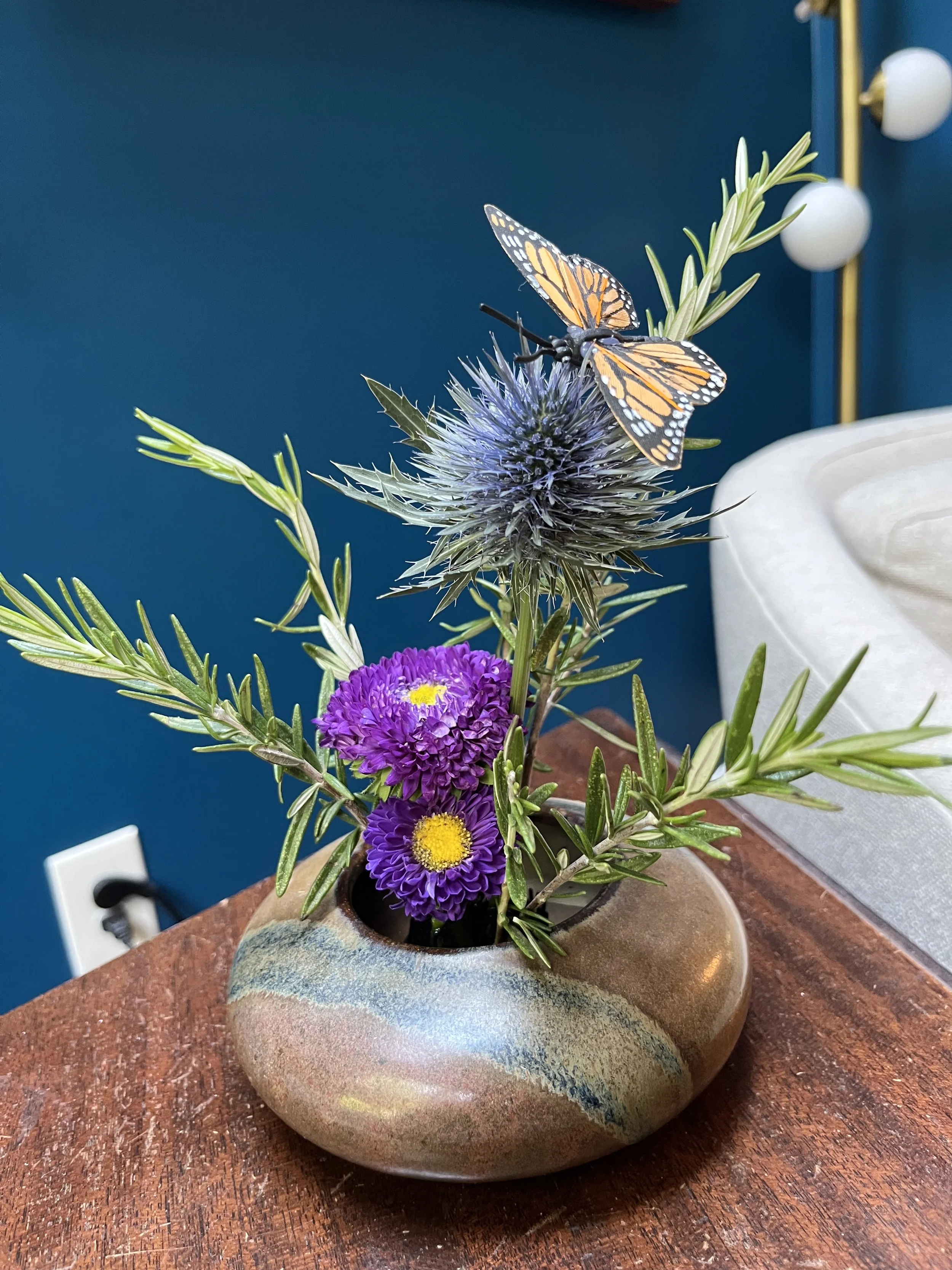 This tiny floral arrangement sits in a ceramic vase and includes purple flowers, rosemary and an orange butterfly.
