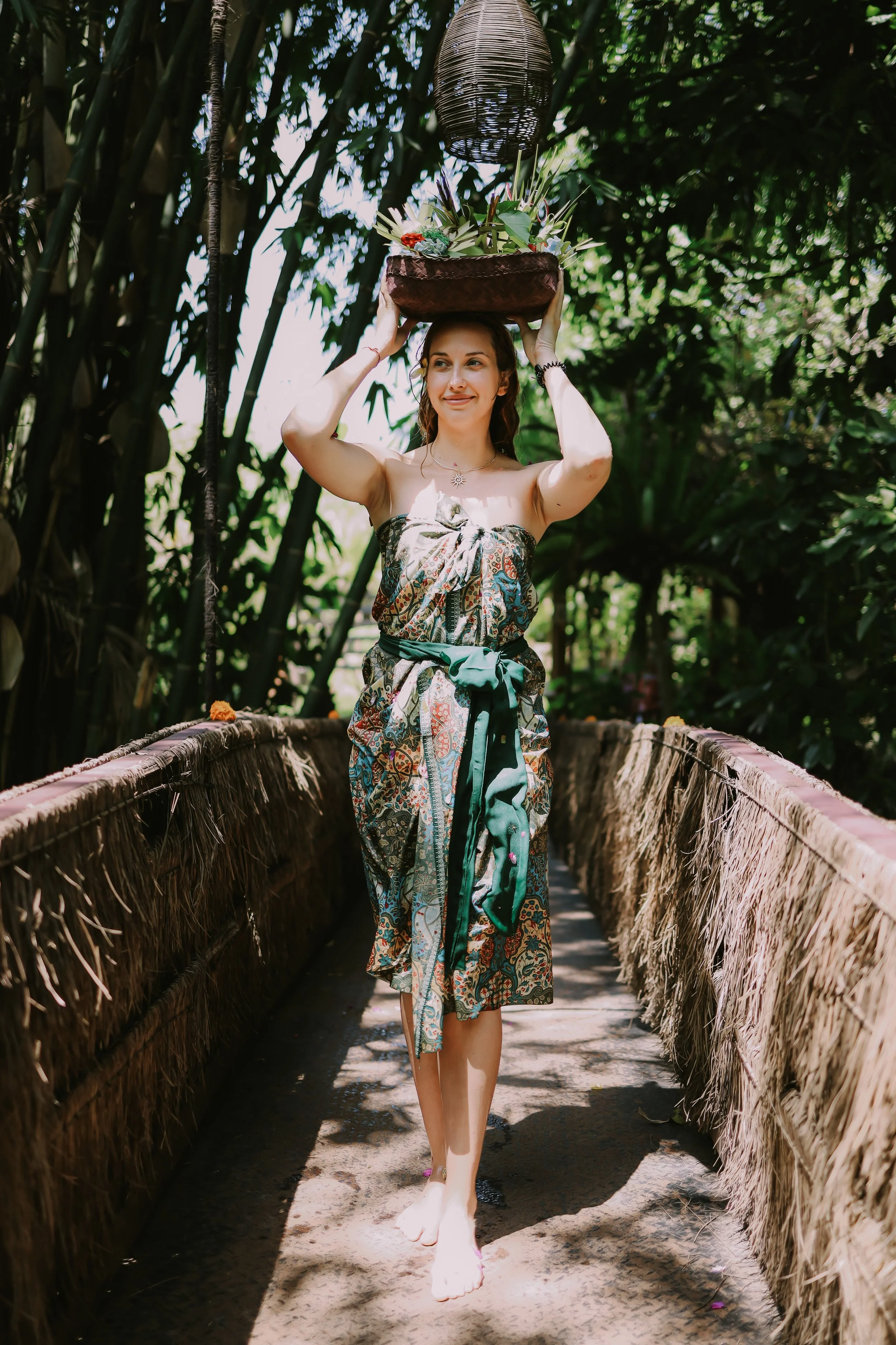 A woman walking on a bamboo pathway in a tropical setting, balancing a large basket of greenery and flowers on her head. She is dressed in a colorful wrap-around skirt and a strapless top, smiling gently.