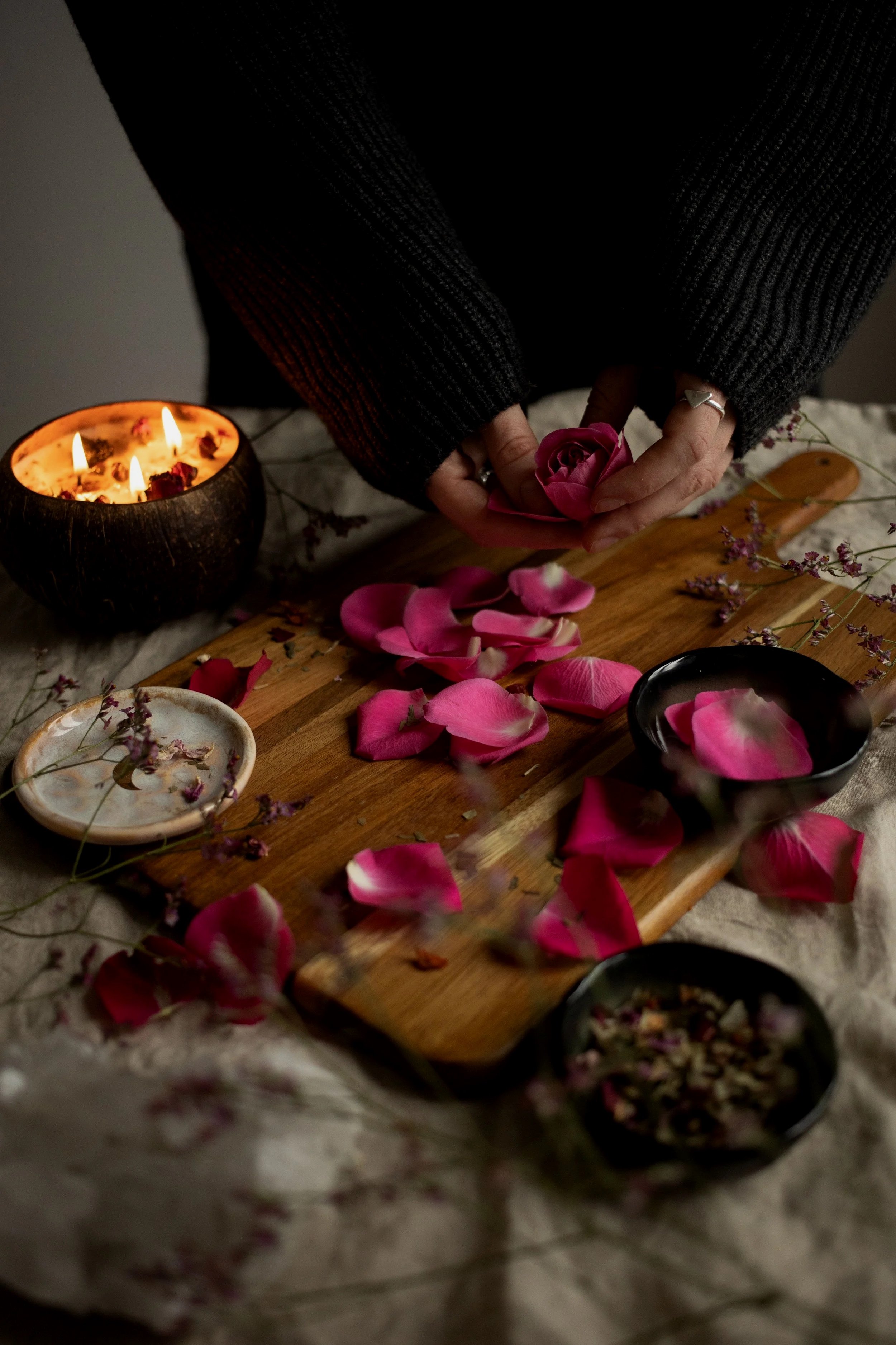 A person in a dark sweater holding a pink rose, surrounded by scattered rose petals and small flowers on a wooden board, with a lit candle nearby illuminating the scene.