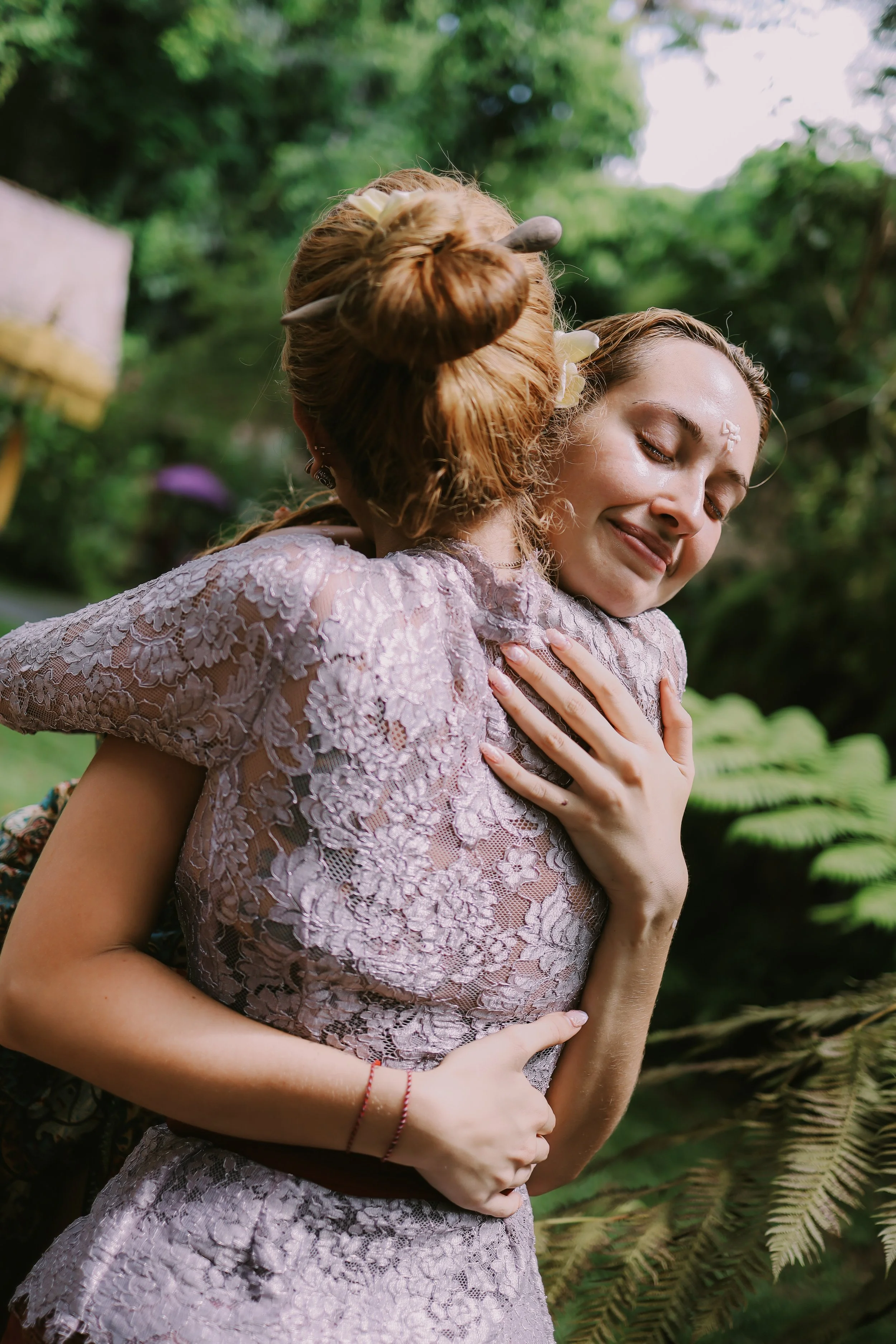 A woman with closed eyes and a gentle smile embraces a woman with her back to the camera, both wearing lace dresses, in a lush outdoor setting with greenery and fern leaves in the background.