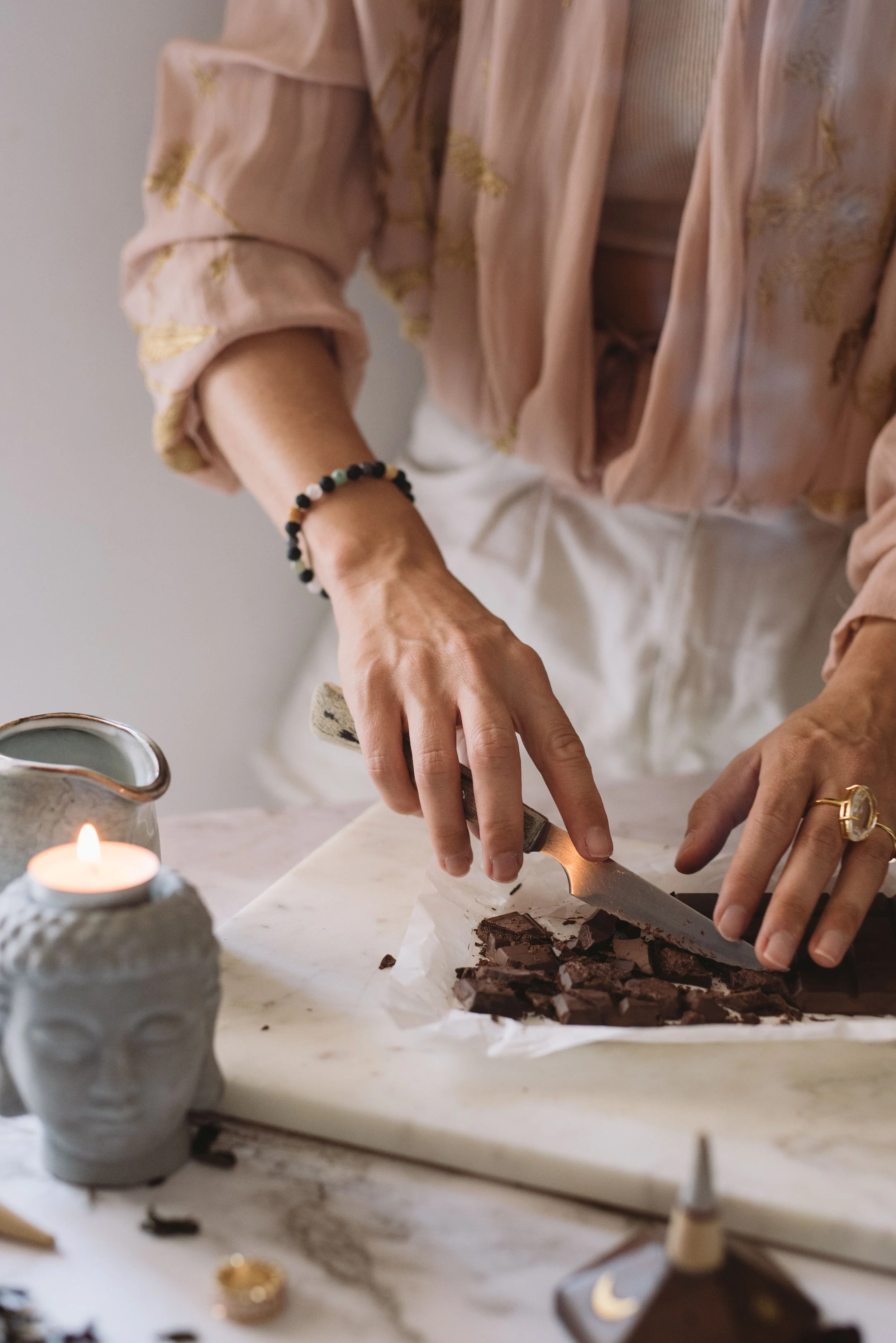 Person slicing a chocolate bar on a white surface, with a lit candle in a small ceramic holder and other jewelry objects on the table.