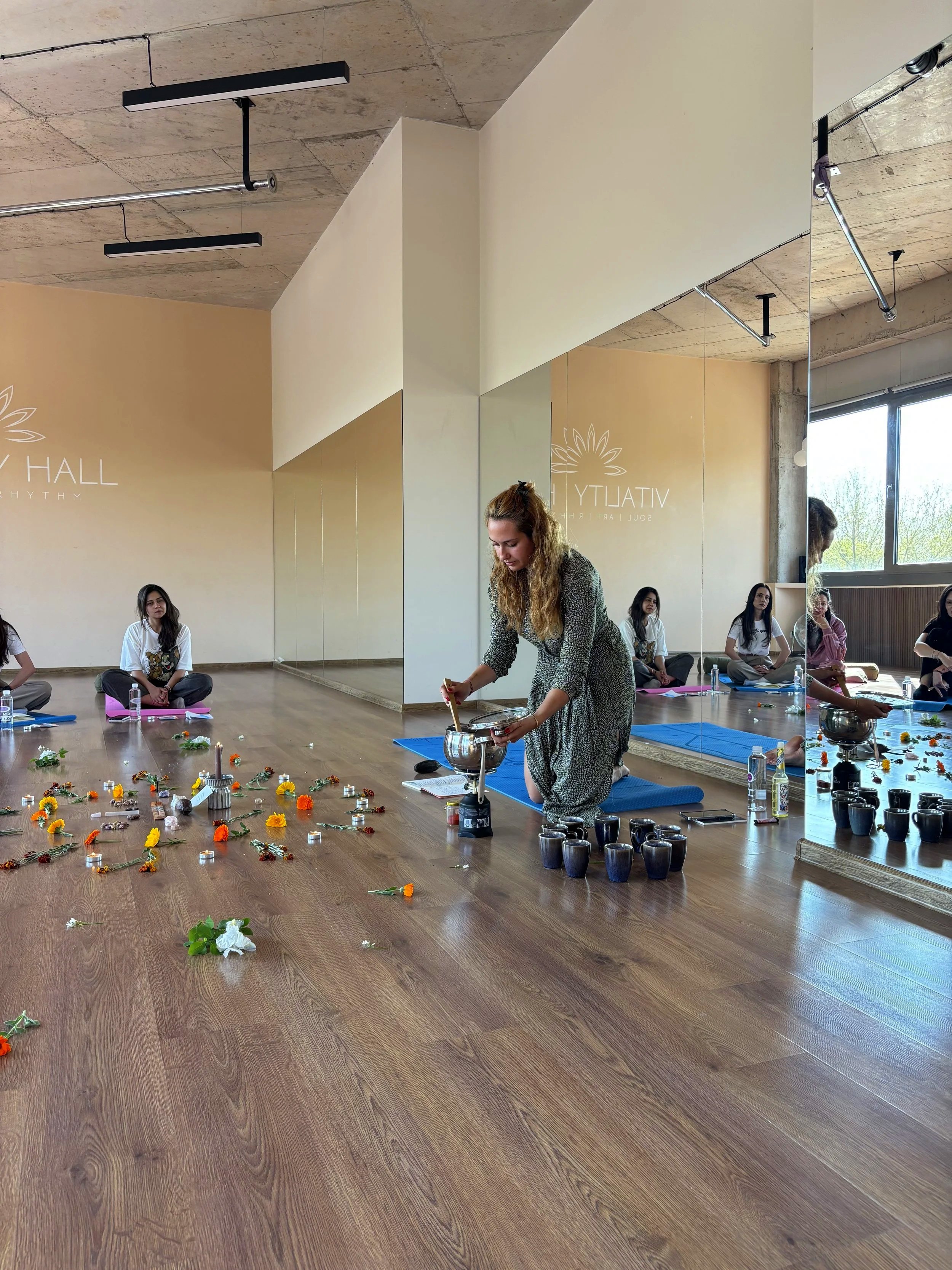 A woman participating in a yoga or meditation class inside a room with large windows and a wood floor. Several women sit cross-legged on yoga mats, some with water bottles nearby. The woman in front is stirring a bowl on a small stove, with flowers and candles arranged on the floor around her.
