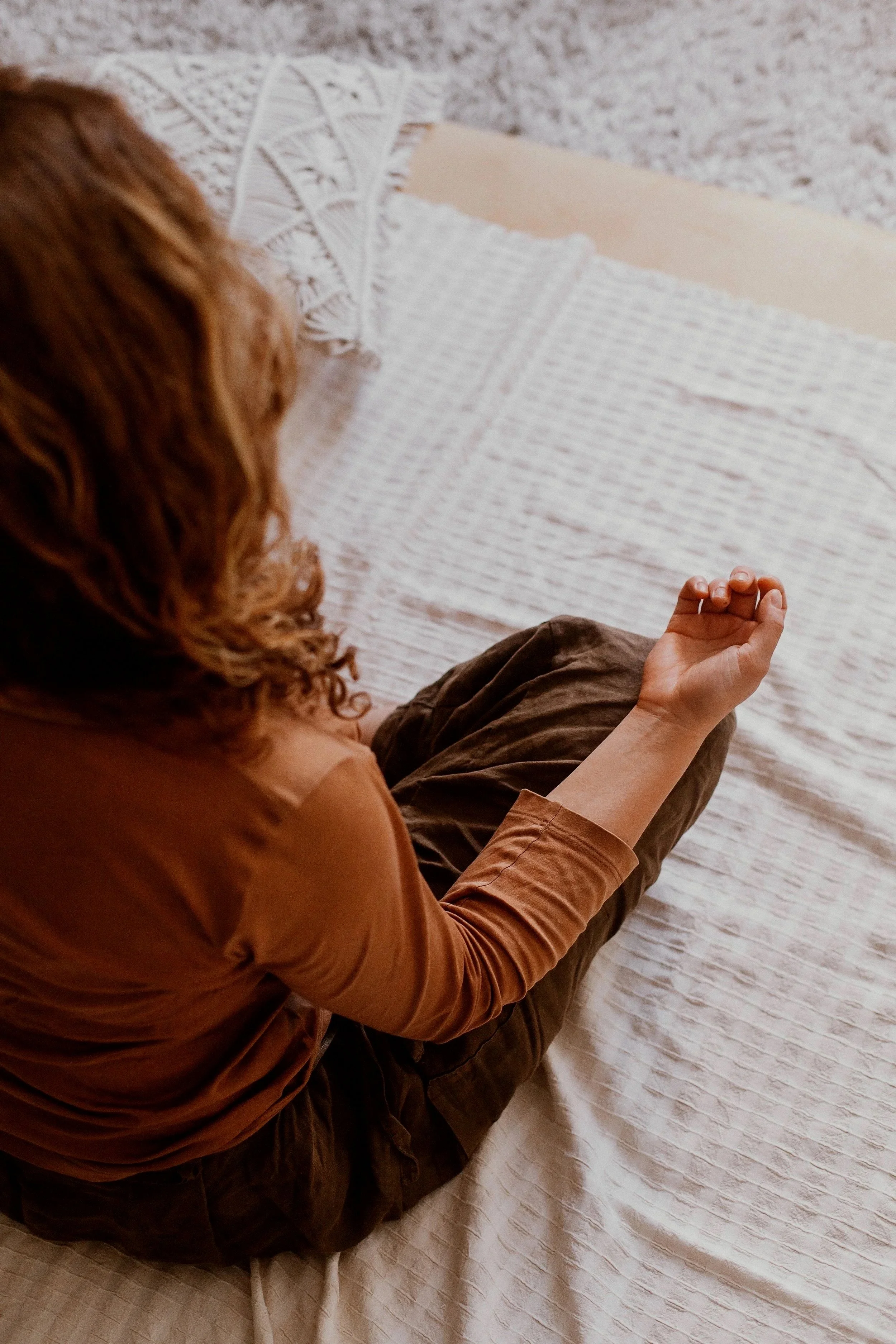 A woman with curly red hair and wearing a brown jacket is sitting on a bed with a beige textured blanket, her legs bent and hands resting on her knee.