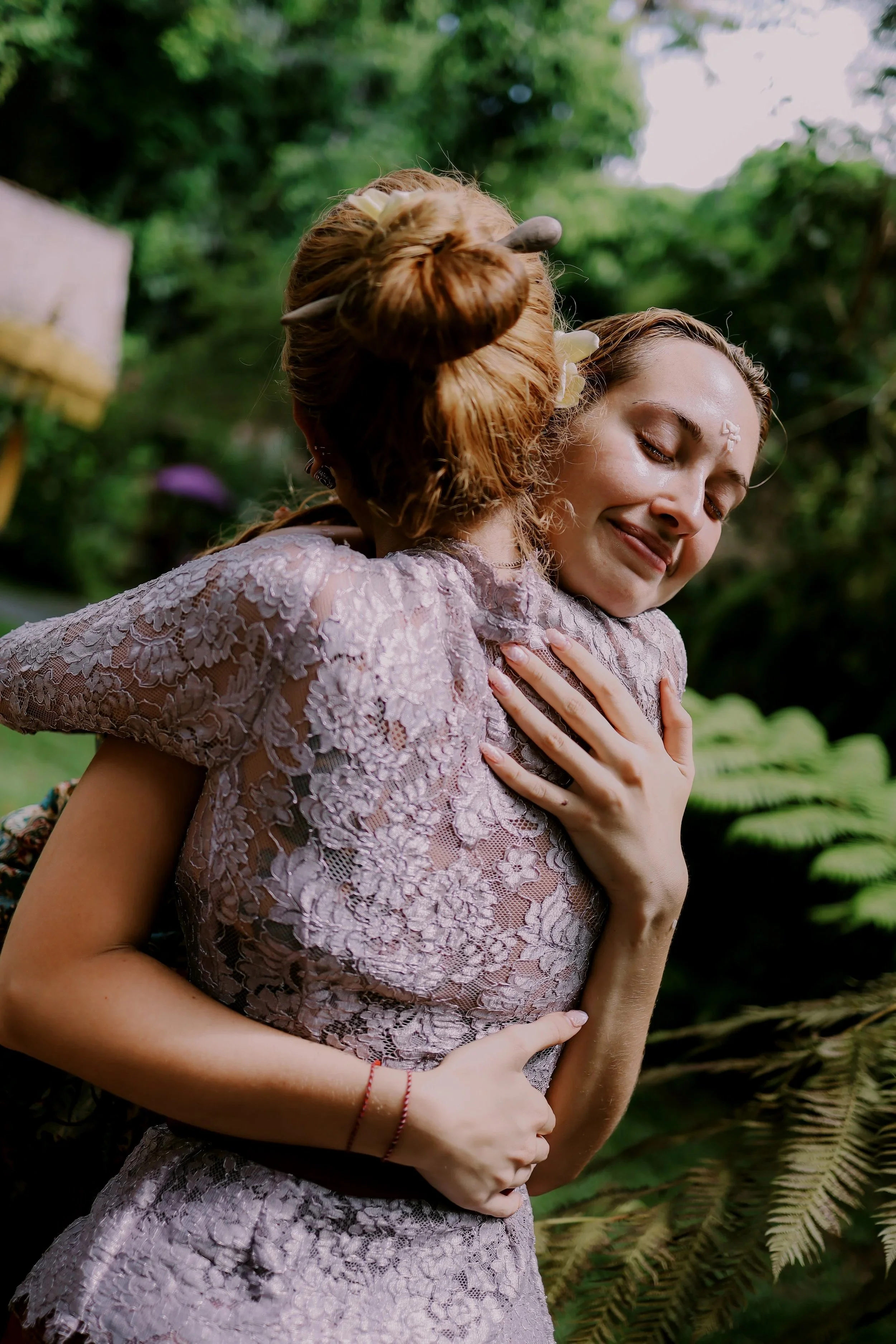 Two women embrace each other outdoors, wearing lace dresses, with lush green foliage in the background.