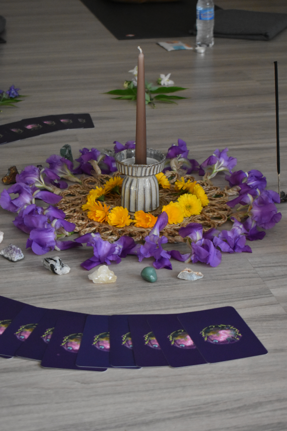 Decorative table with a central candle surrounded by purple flowers, yellow flowers, crystals, and tarot cards.