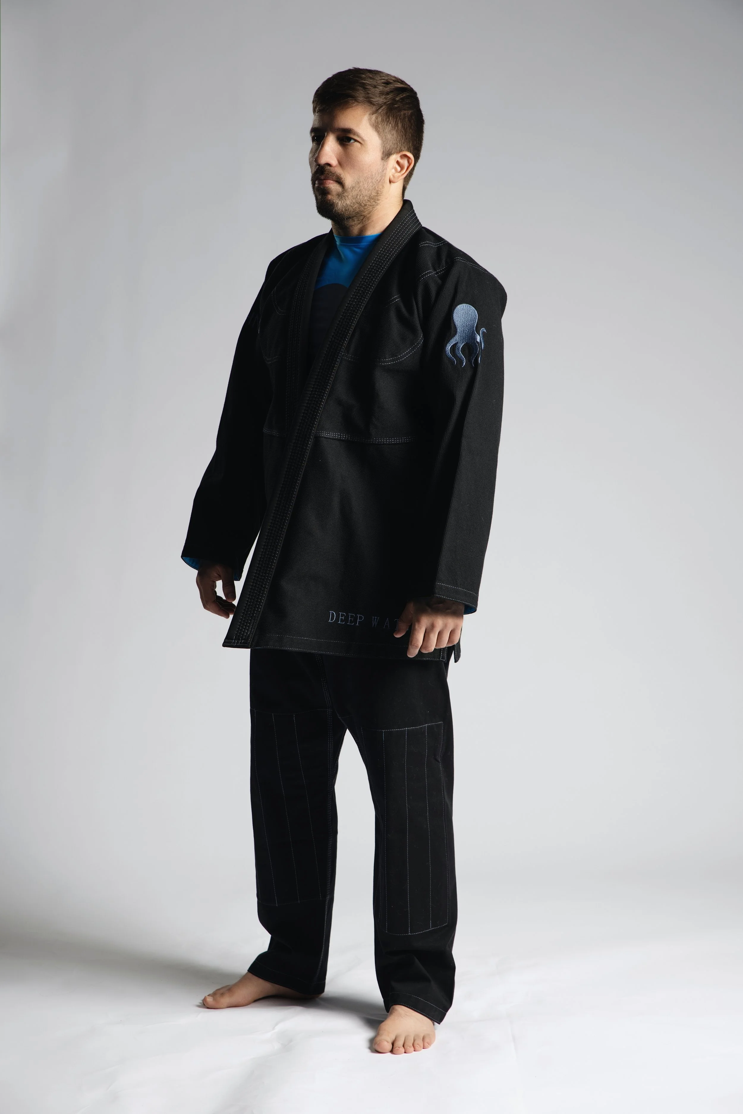 A man standing barefoot against a plain background, wearing a black martial arts gi with a blue octopus patch on the sleeve and the words 'Deep Water' embroidered on the belt.