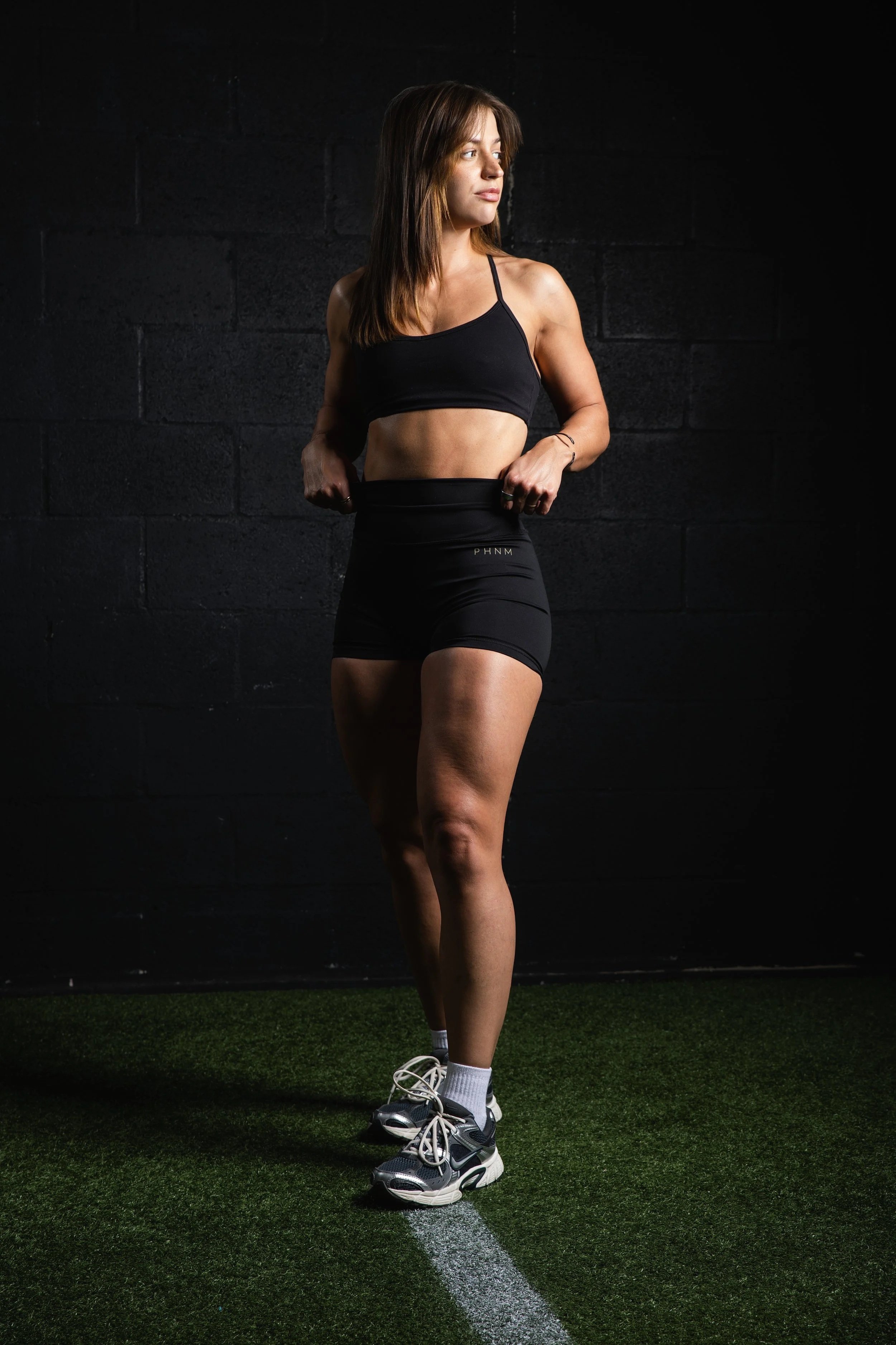 A woman in athletic clothing standing on a sports field, adjusting her waistband, with a dark background and bright lighting highlighting her and her workout gear.