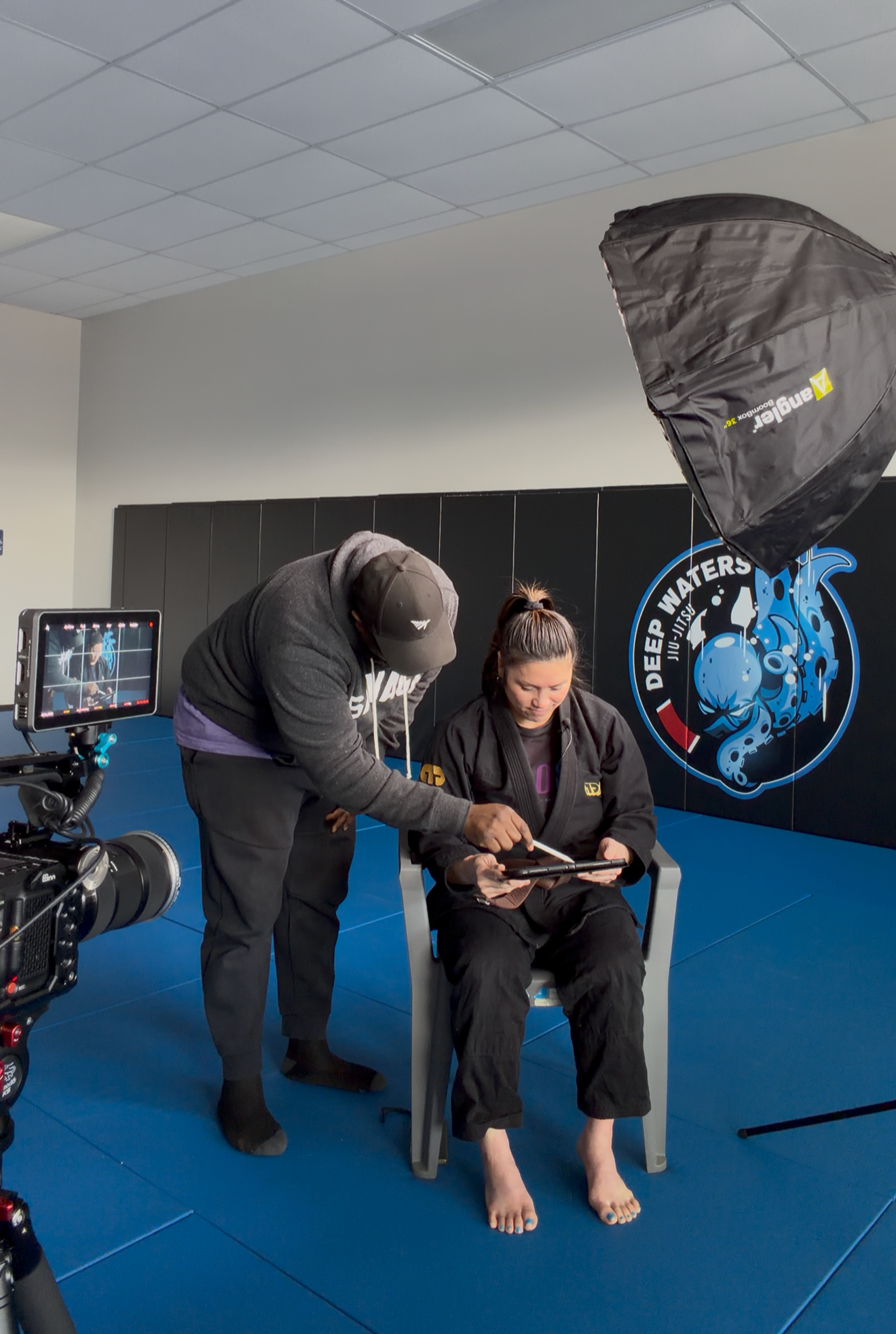 Two people are filming a woman sitting in a chair with her feet on the floor in a martial arts gym. The woman is wearing a gi and appears to be preparing for a martial arts demonstration or training session. There is professional photography equipment, including a softbox and a camera, in the scene.