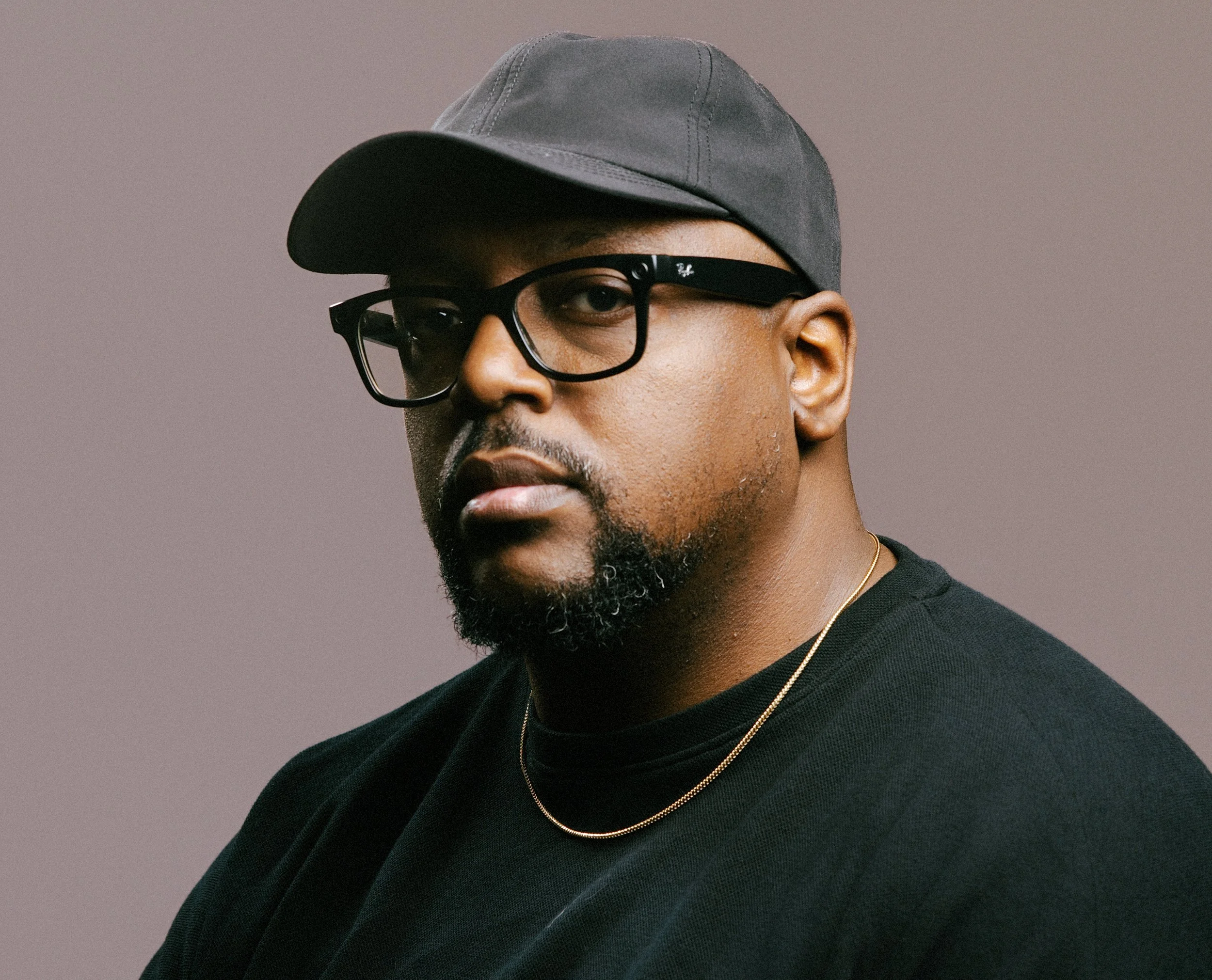 A man with a dark beard and mustache wearing a gray baseball cap, black glasses, a black shirt, and two gold necklaces against a gray background.