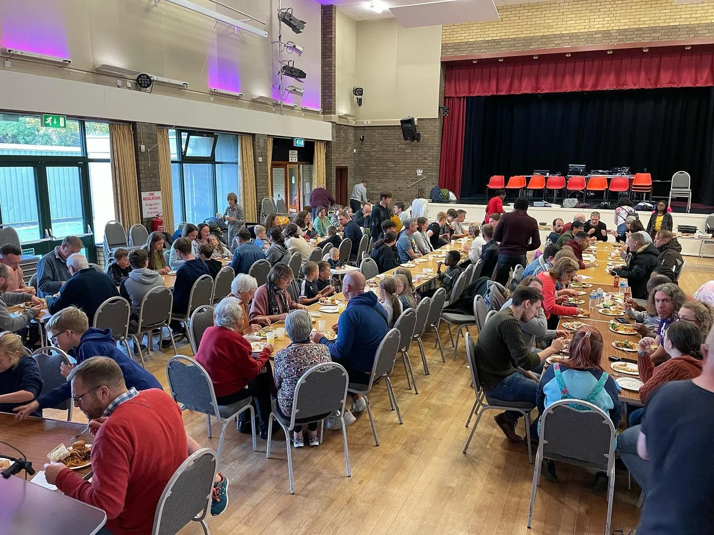 Large indoor community hall filled with people sitting at long tables, eating a Big Sunday Lunch together. Church family.