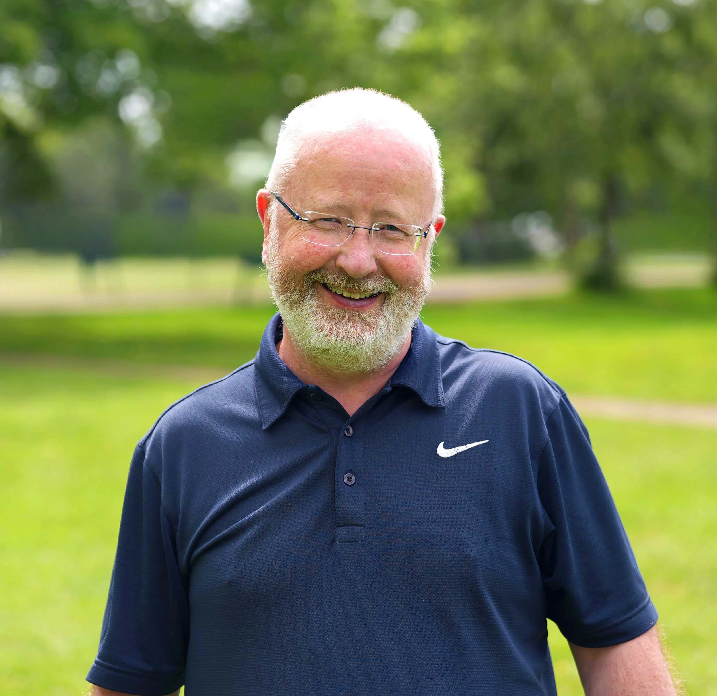 Close-up of a smiling man with white hair and beard wearing glasses and a navy blue polo shirt, outdoors with green trees and grass in the background.