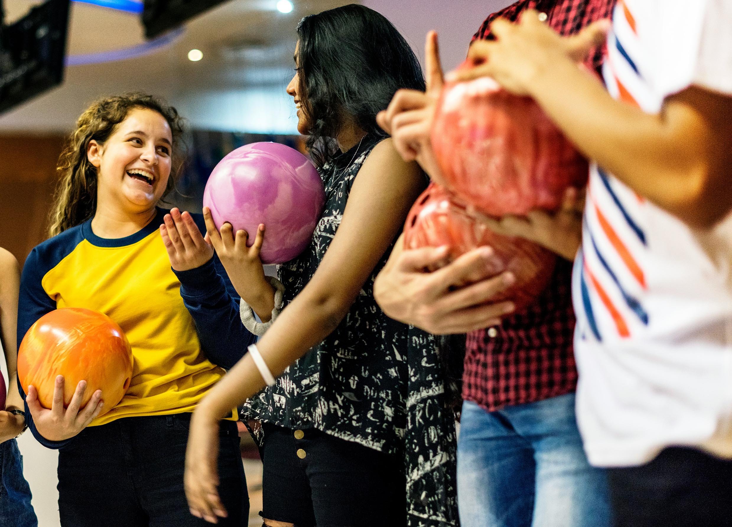 Group of people standing in line, including a girl with curly hair and yellow and blue shirt holding a pumpkin, and women with dark hair holding balloons, socializing indoors.