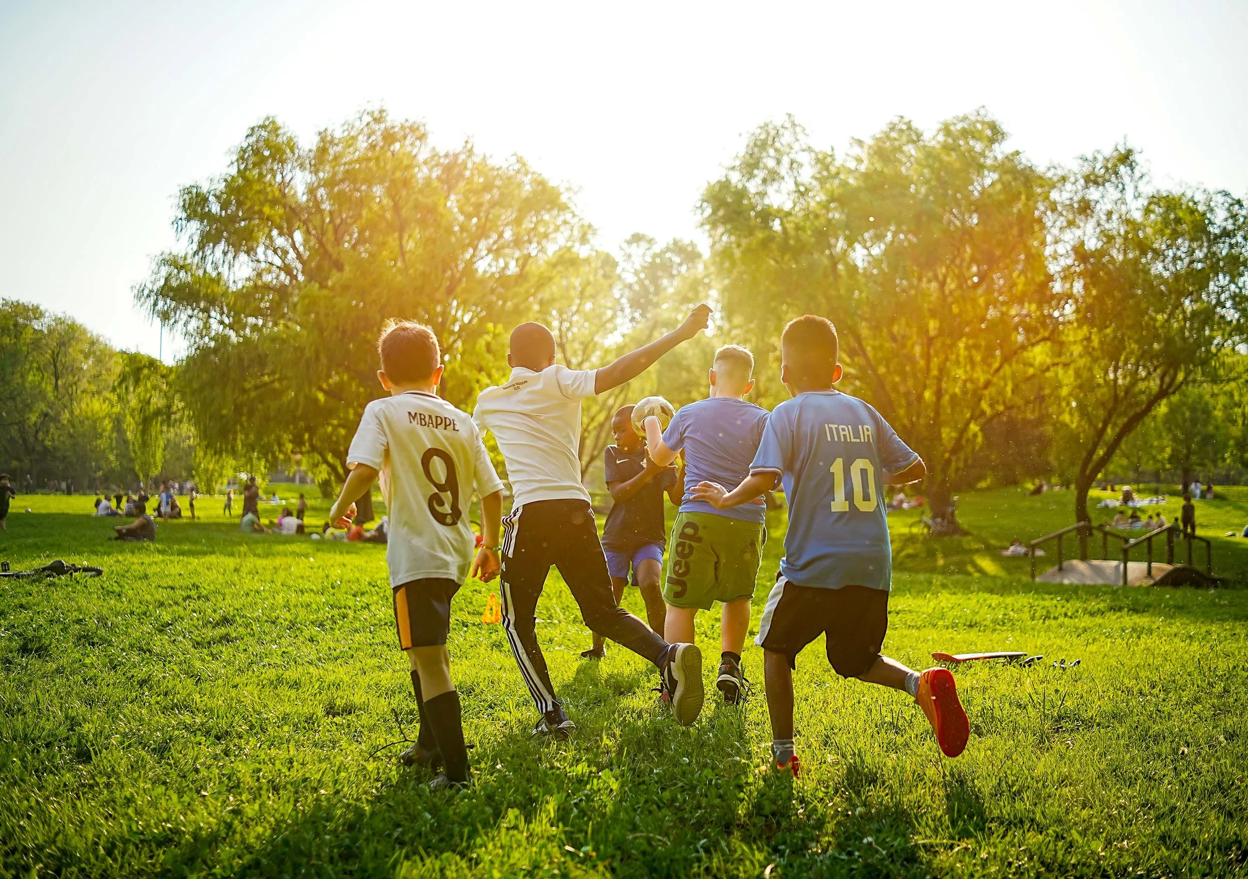 Children playing soccer in a park during sunset, facing away from the camera.