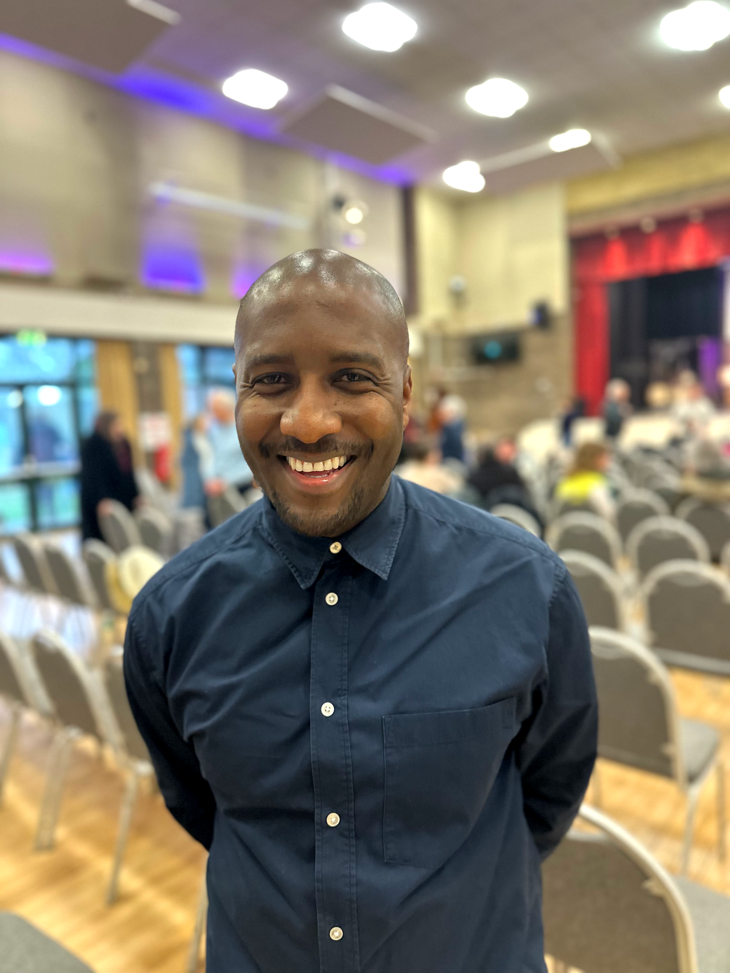 A smiling man with a shaved head wearing a dark blue button-up shirt in a large indoor event space with chairs and people in the background.