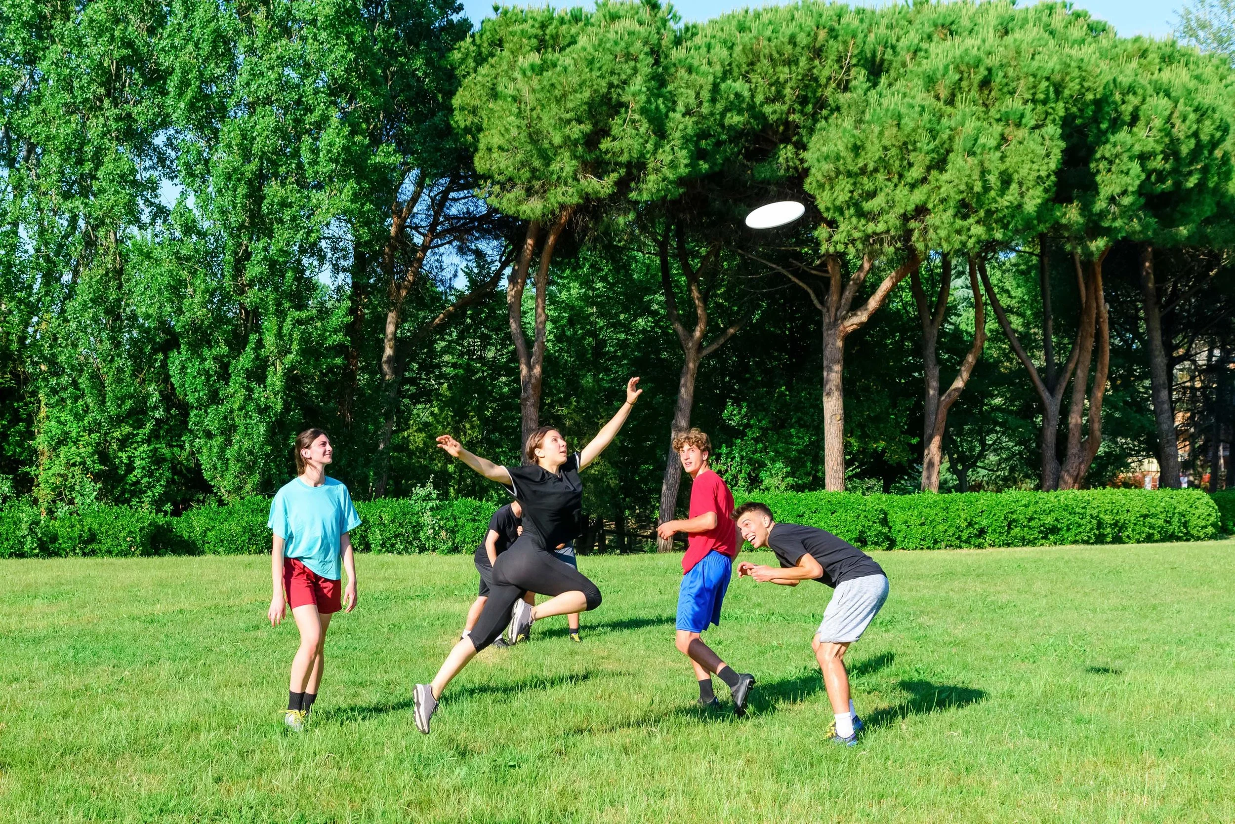 A group of young people playing flag football outdoors on a grassy field surrounded by trees on a sunny day.