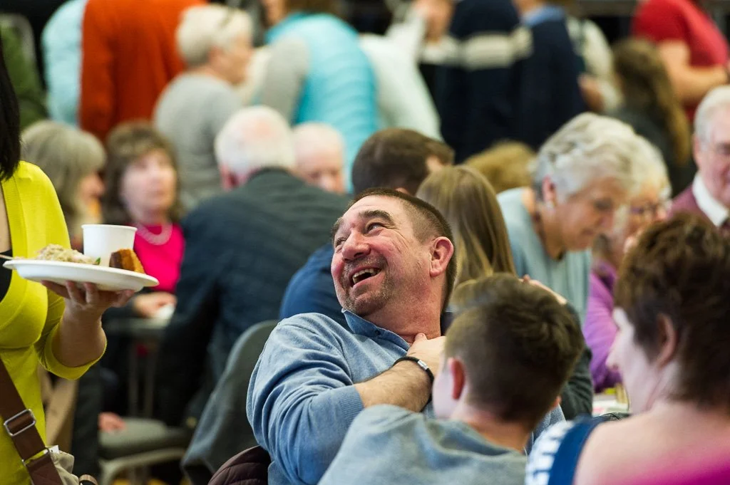 A man laughing at a church gathering with many people seated at tables, with a woman serving food in the foreground.