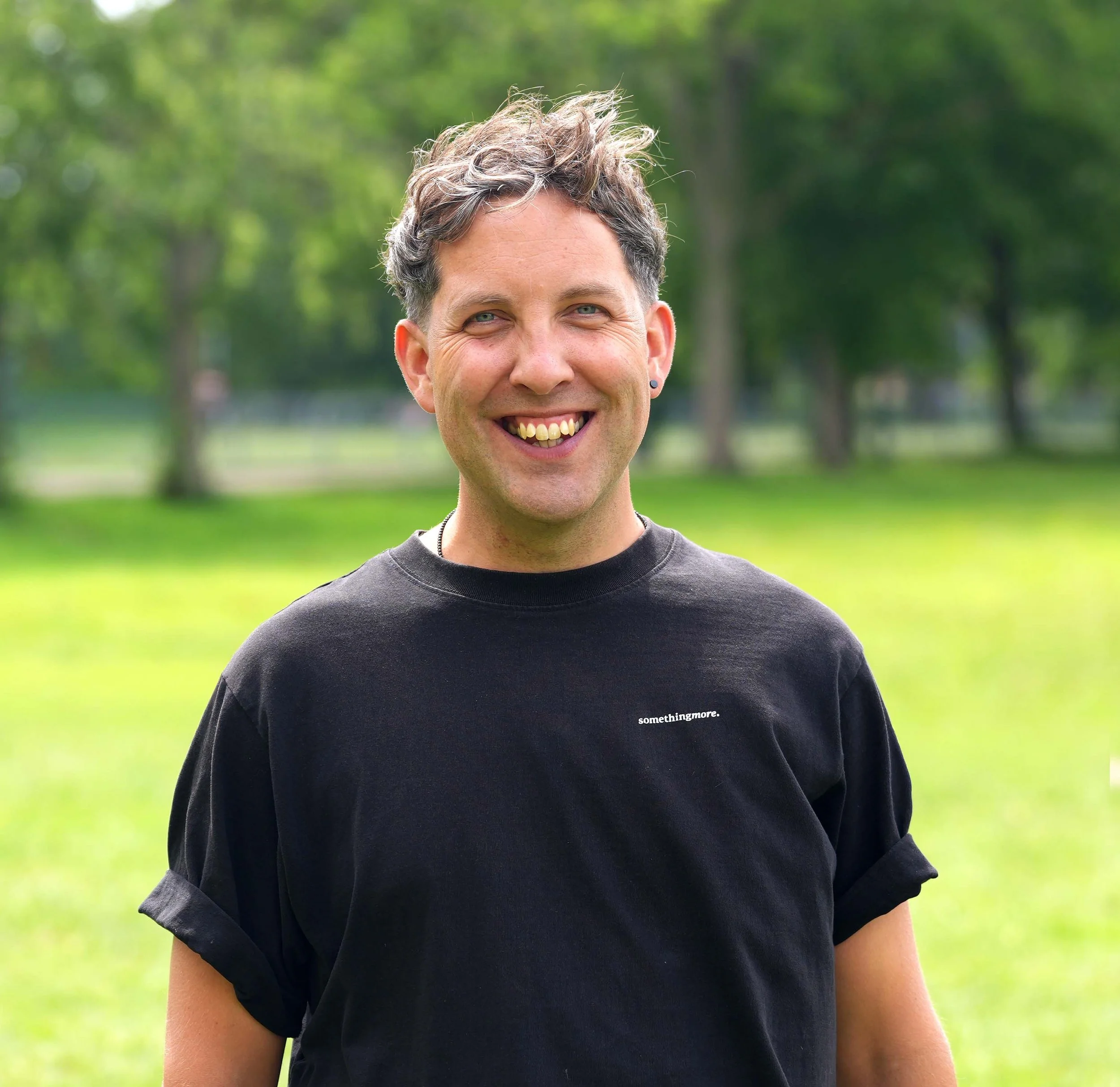 A person smiling outdoors in a park with green trees in the background, wearing a black t-shirt with the word 'somethingmore' on it.