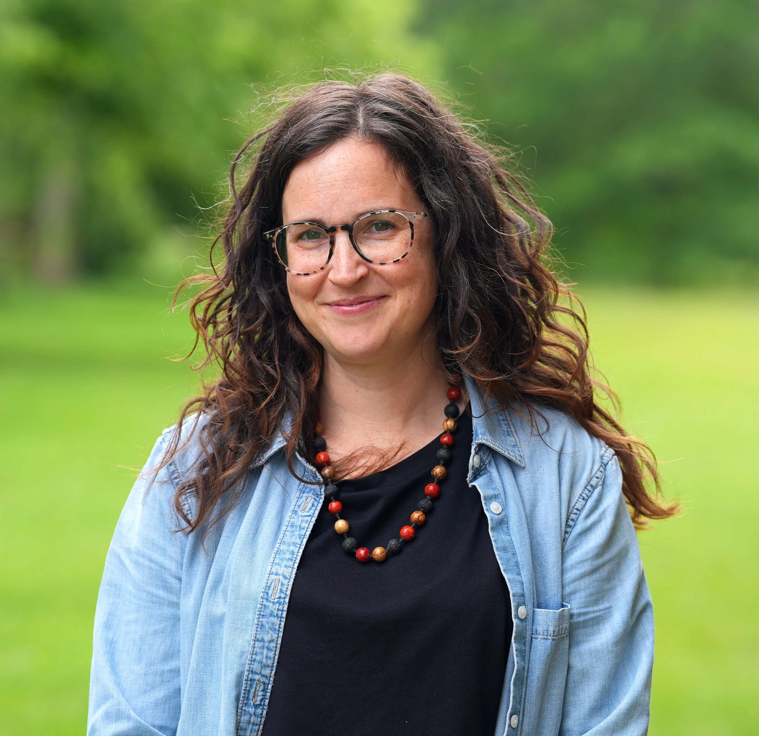 A woman with curly brown hair wearing glasses, a black shirt, and a blue denim jacket, standing outdoors with a green blurred background.