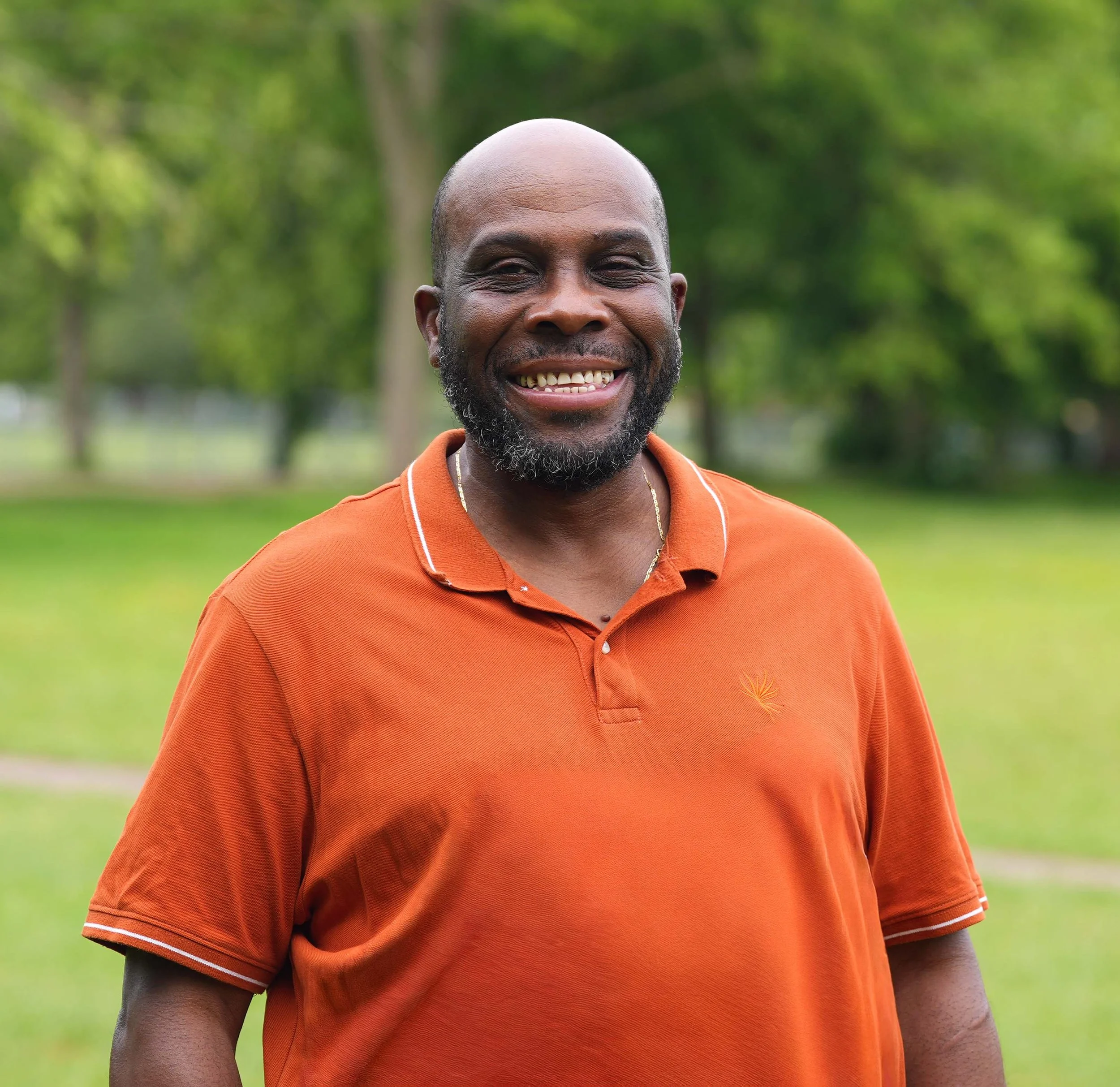 Close-up of a smiling man with dark skin, a beard, wearing an orange polo shirt with white trim, standing outdoors in a park with green trees and grass in the background.