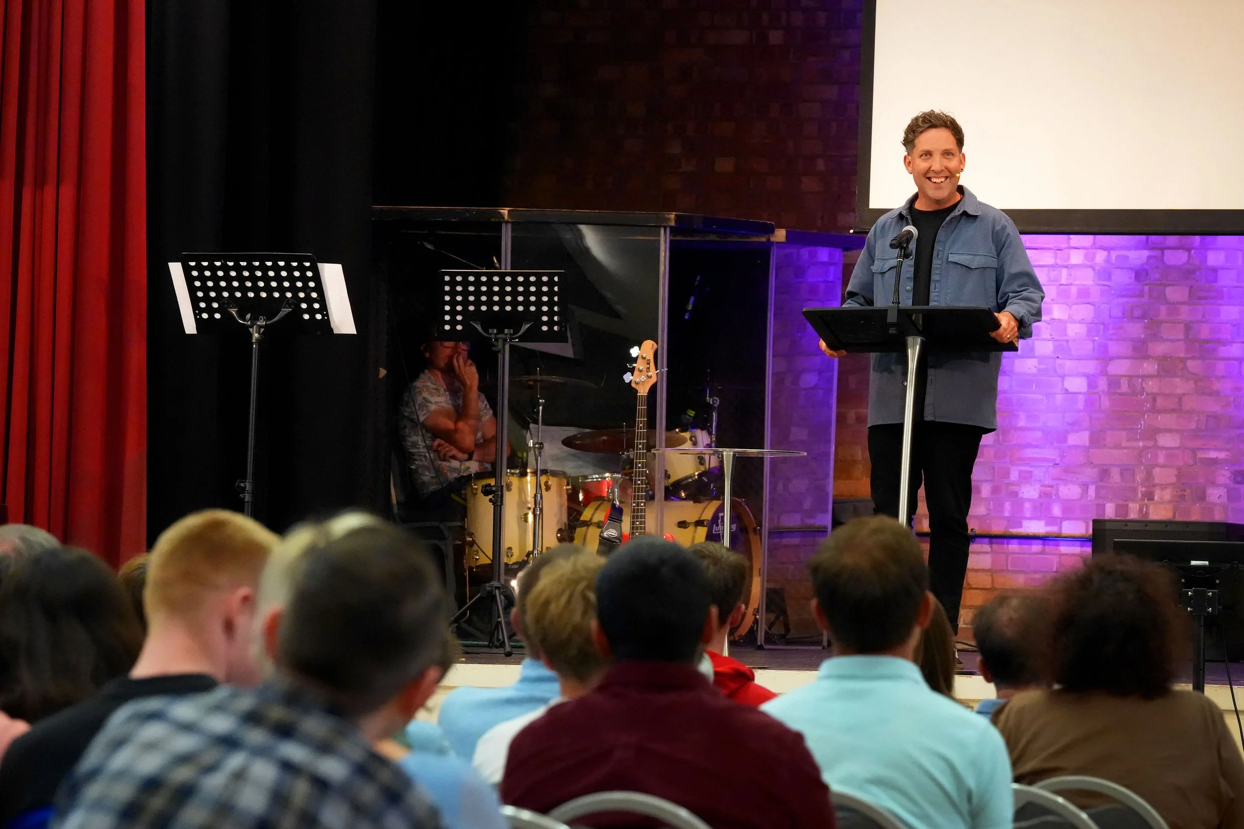 A man is speaking at a podium in front of an audience. Behind him, a woman and a drum set are visible on stage. #Grace #Church #SundayServices