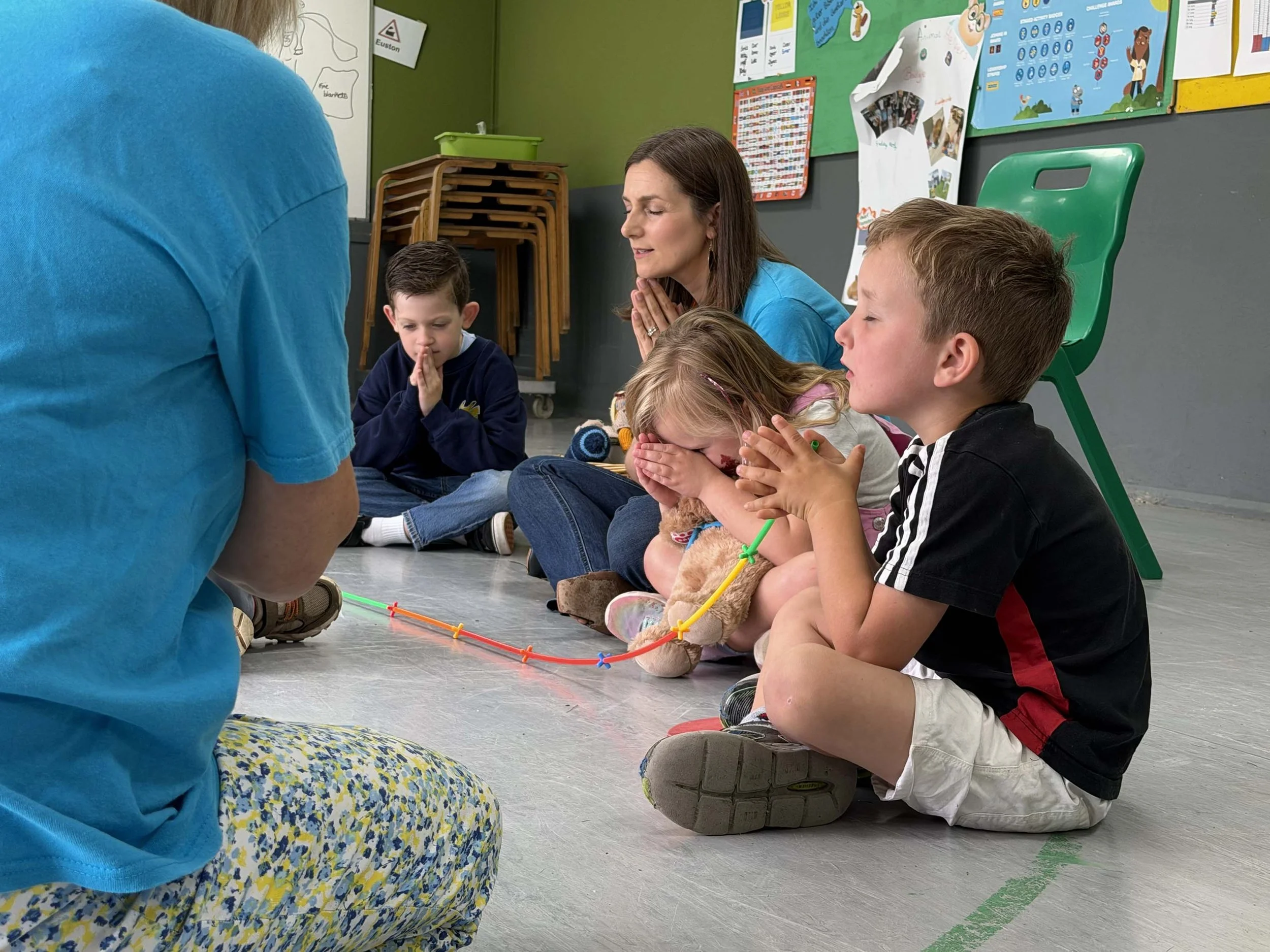 Children sitting cross-legged on the floor in a classroom engaging in a prayer or meditation led by an adult.
