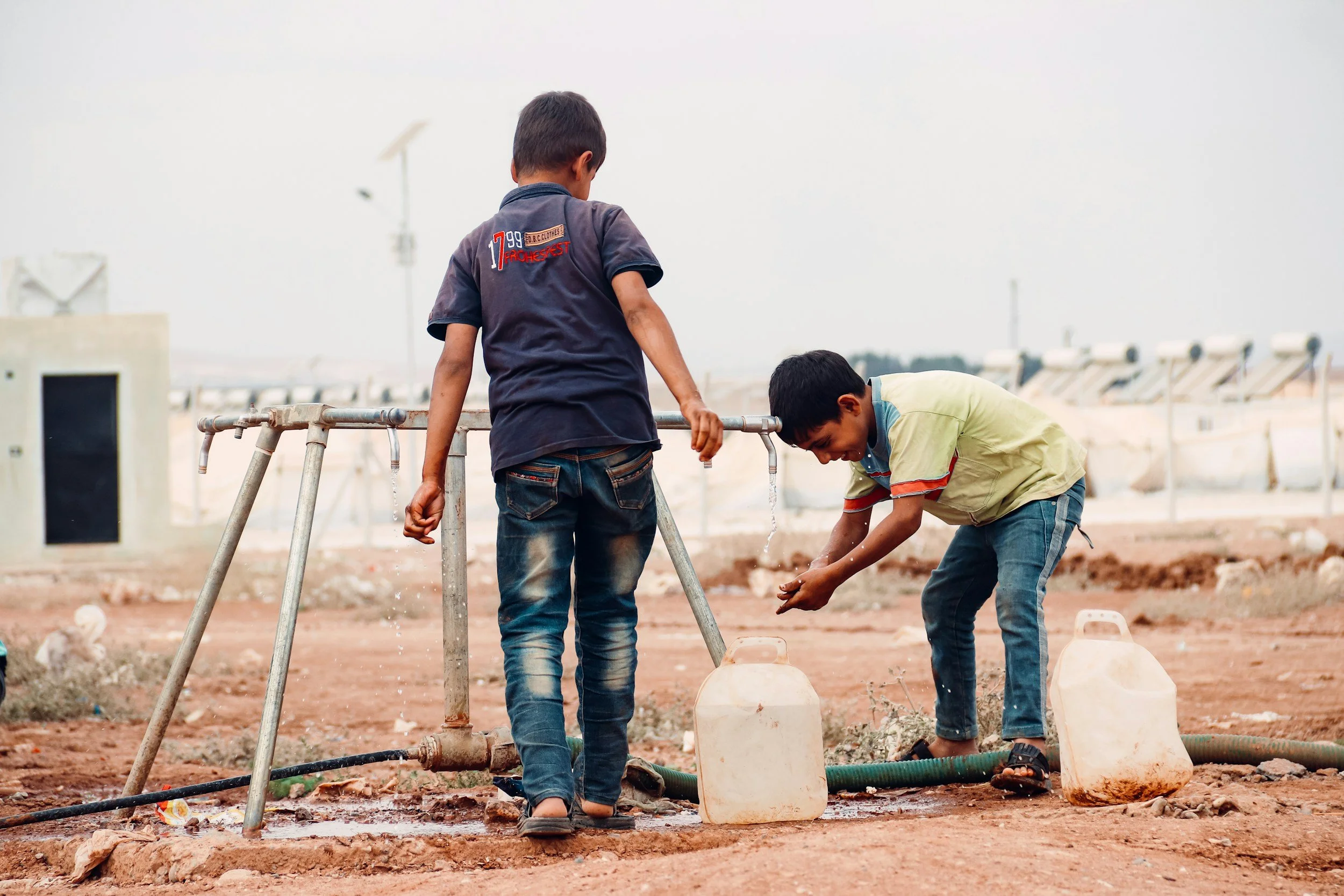 Two young boys playing with water from a pipe on a construction site or open area, surrounded by containers and hoses.
