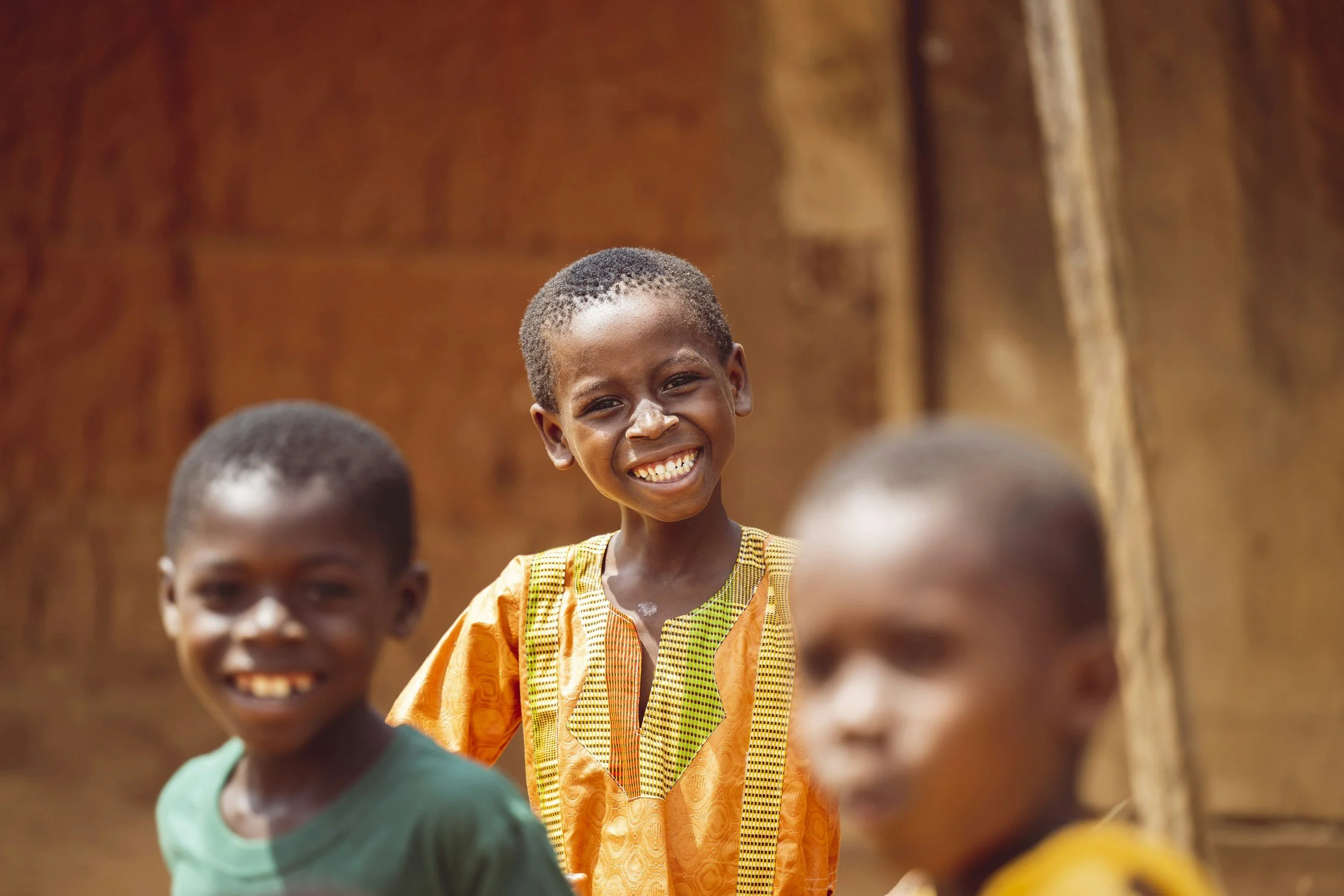 A smiling young girl with a short hairstyle, wearing a colorful traditional dress, surrounded by other children in a warm earth-toned outdoor setting.