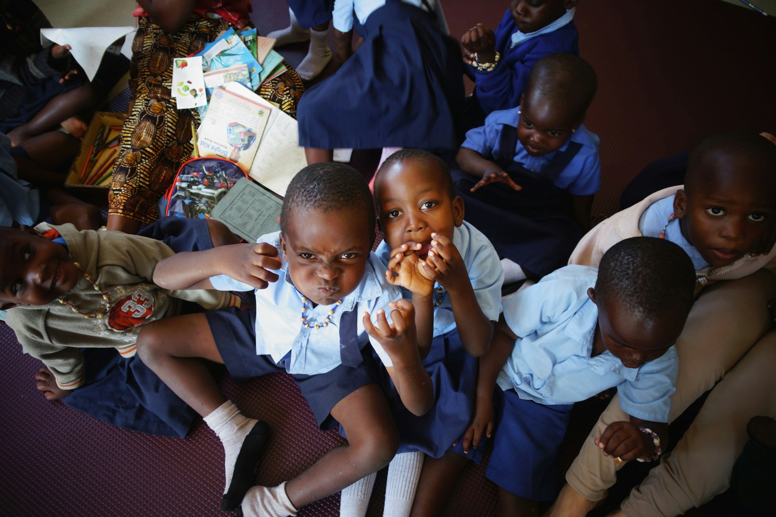 A group of children sitting on a maroon mat, looking up at the camera. They are wearing school uniforms in blue and beige, with some wearing necklaces and bracelets. There are books and drawings scattered around them.
