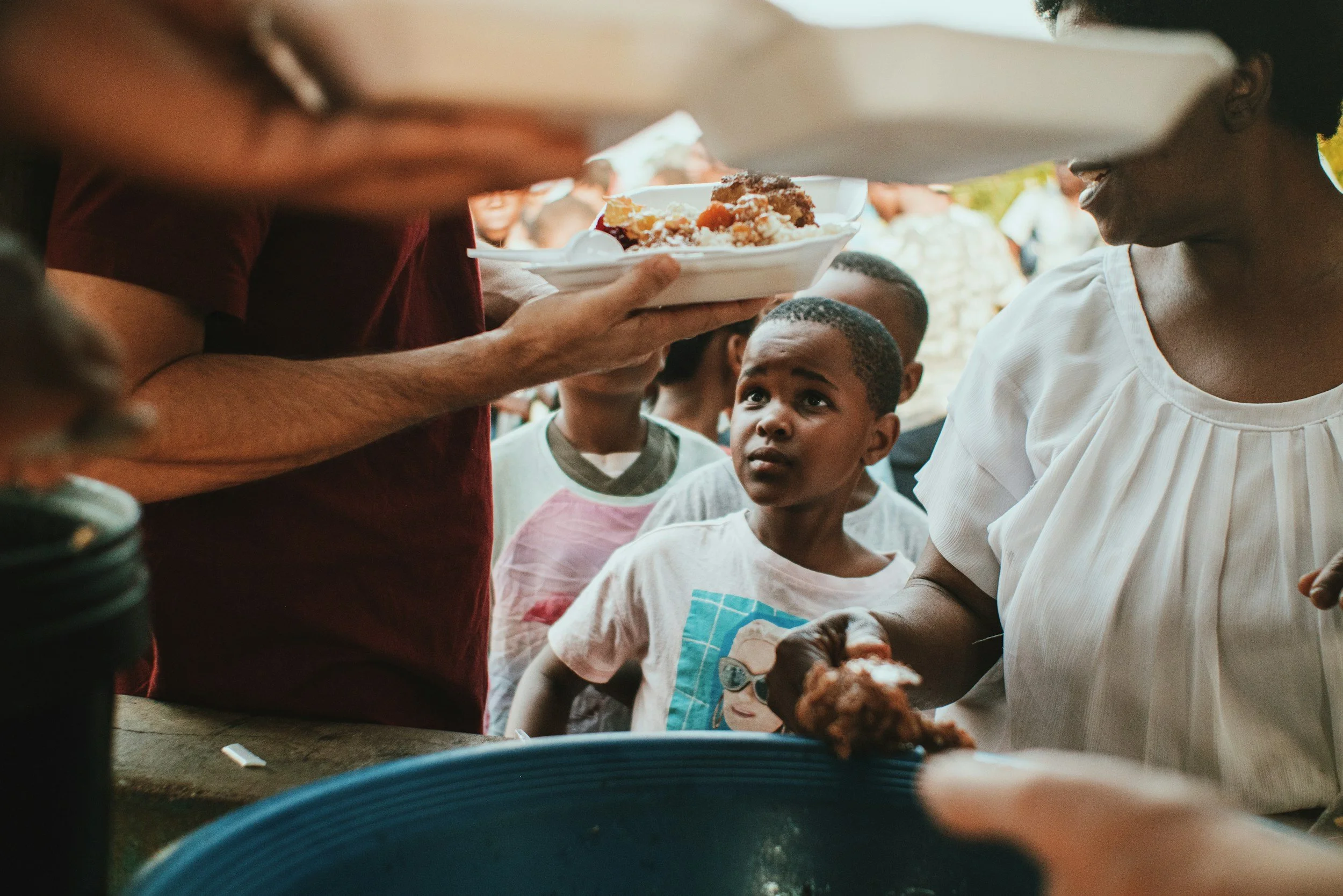 A crowd of people gathered outdoors, with a focus on a young boy looking up as someone offers a plate of food. The scene suggests a communal meal or food distribution event.