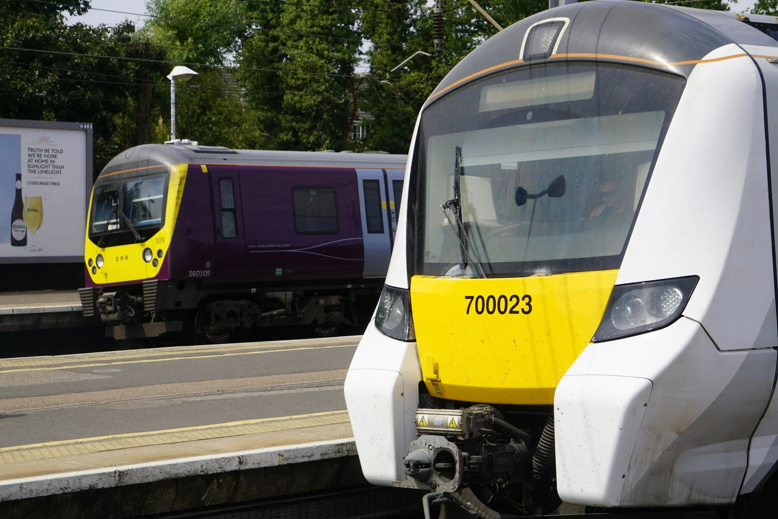 Close-up of two modern trains on parallel tracks, one white with yellow accents in the foreground, and a purple and yellow train in the background, with green leafy trees and a billboard in the distance.