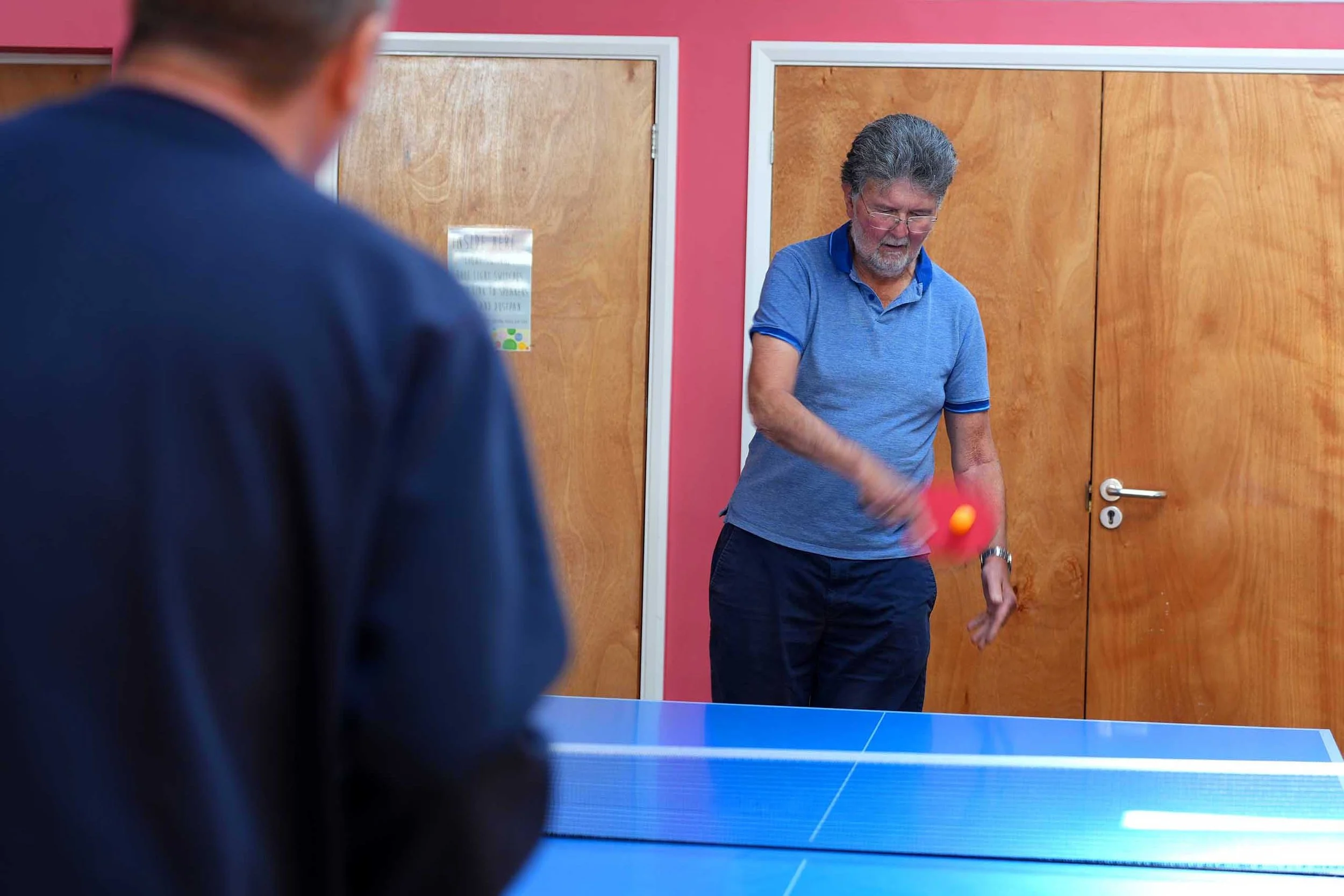 A man with glasses and a beard wearing a blue polo shirt playing ping pong, with another person whose back is to the camera, in a room with pink and wooden doors. The Shed, a group for men in Kempston.