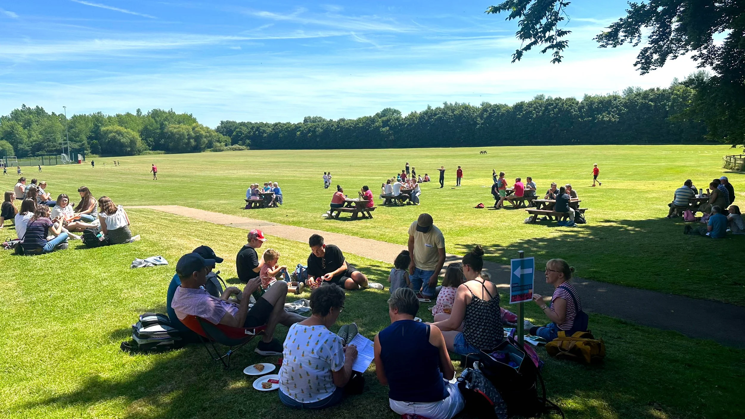People gathered outdoors on a sunny day in a park-like setting, sitting on grass and picnic tables, socializing and enjoying food near an open field with trees in the background. #Community #Church #Bedford
