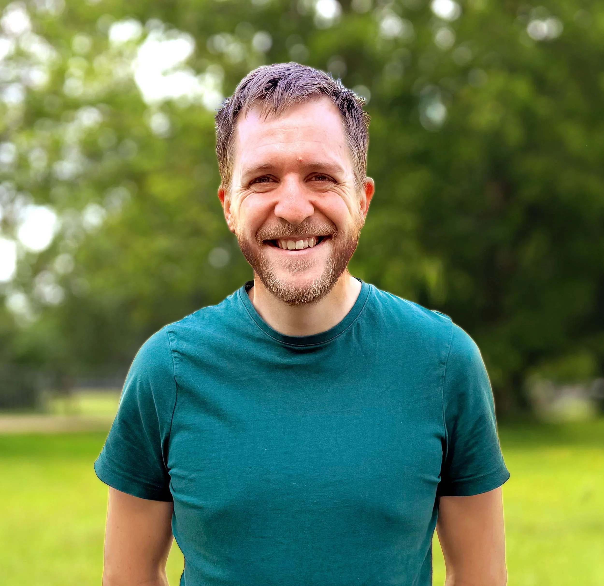 Close-up of a smiling man with short brown hair, beard, wearing a teal t-shirt, outdoors with green trees in the background.