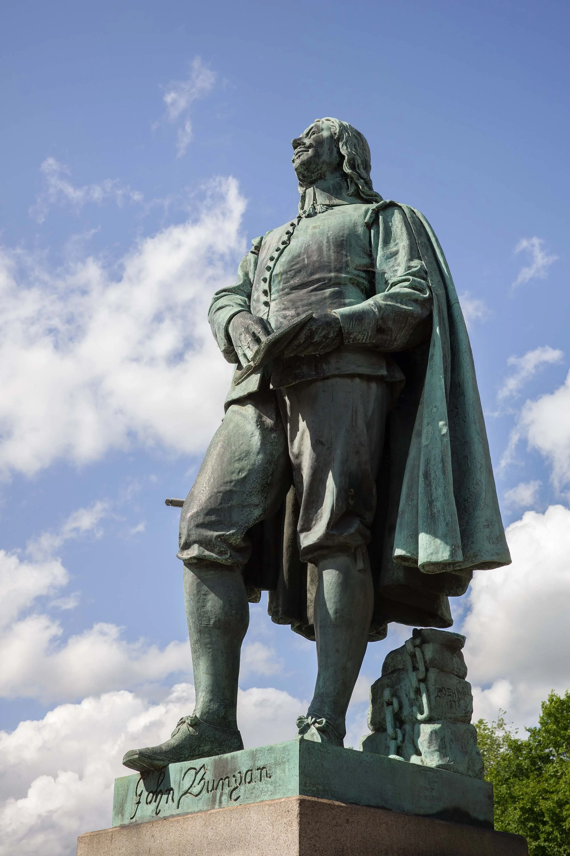 Statue of a historical man standing with one foot slightly forward, holding a book in one hand and a shield or similar object in the other, with an inscription reading 'Joan Bunyan' on the base, set against a partly cloudy sky.