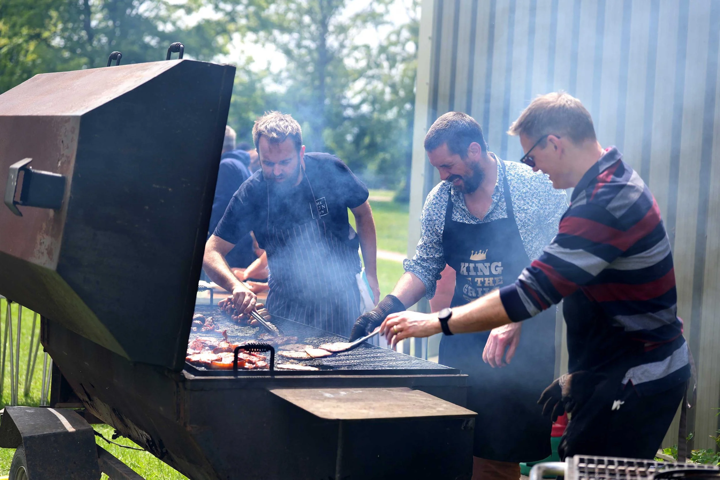 People grilling meat outdoors near a metal building with trees in the background.