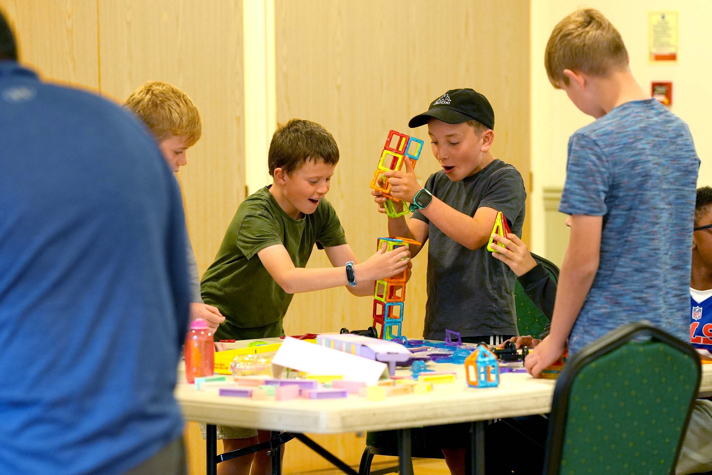 Group of children playing with colorful magnetic building blocks at a table in a room with yellow walls.