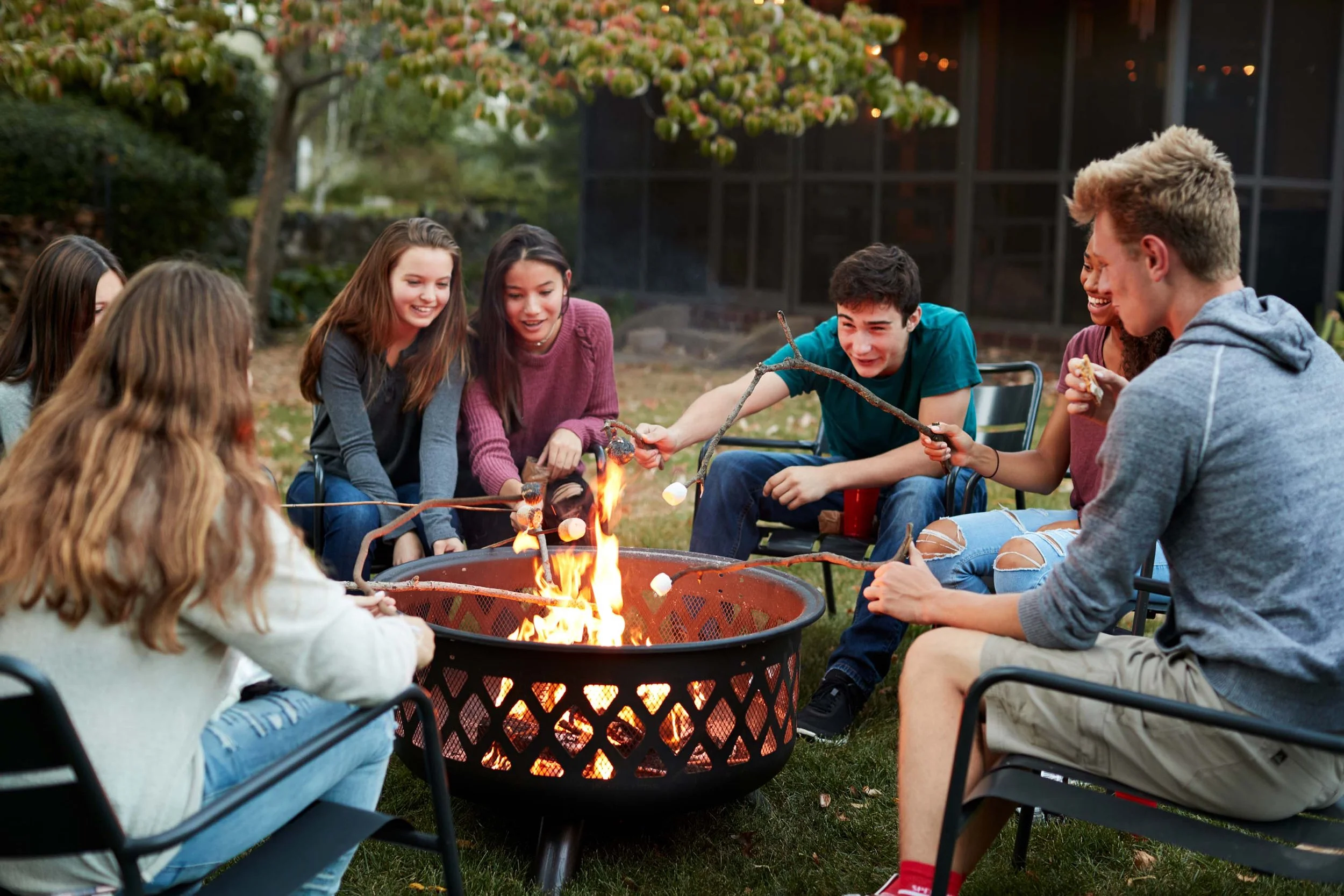 Group of teenagers sitting around a fire pit outdoors, roasting marshmallows on sticks and smiling.