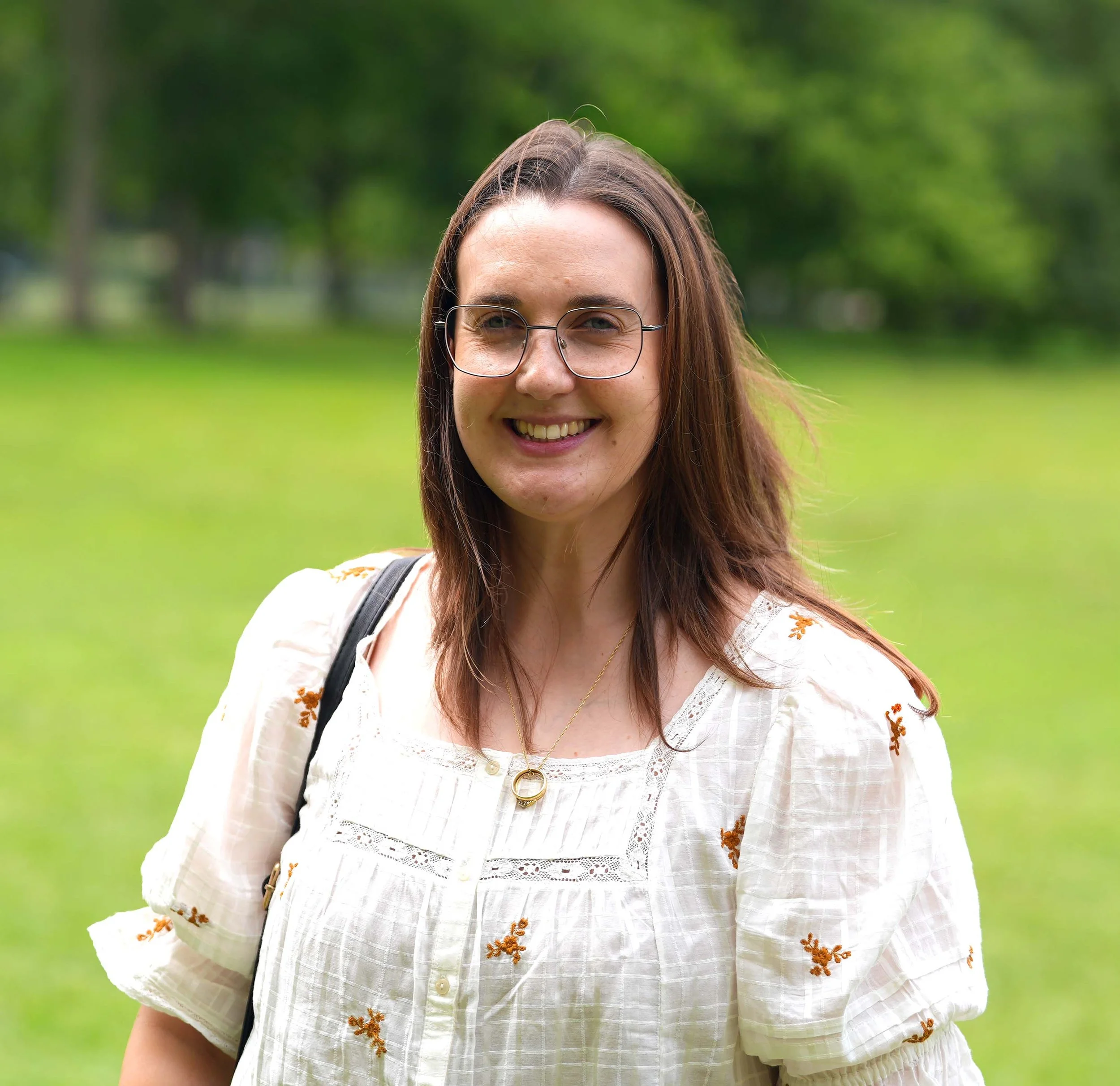 A smiling young woman with long brown hair wearing glasses, a white embroidered blouse, and a gold necklace, standing outdoors in front of a blurred green background of trees and grass.