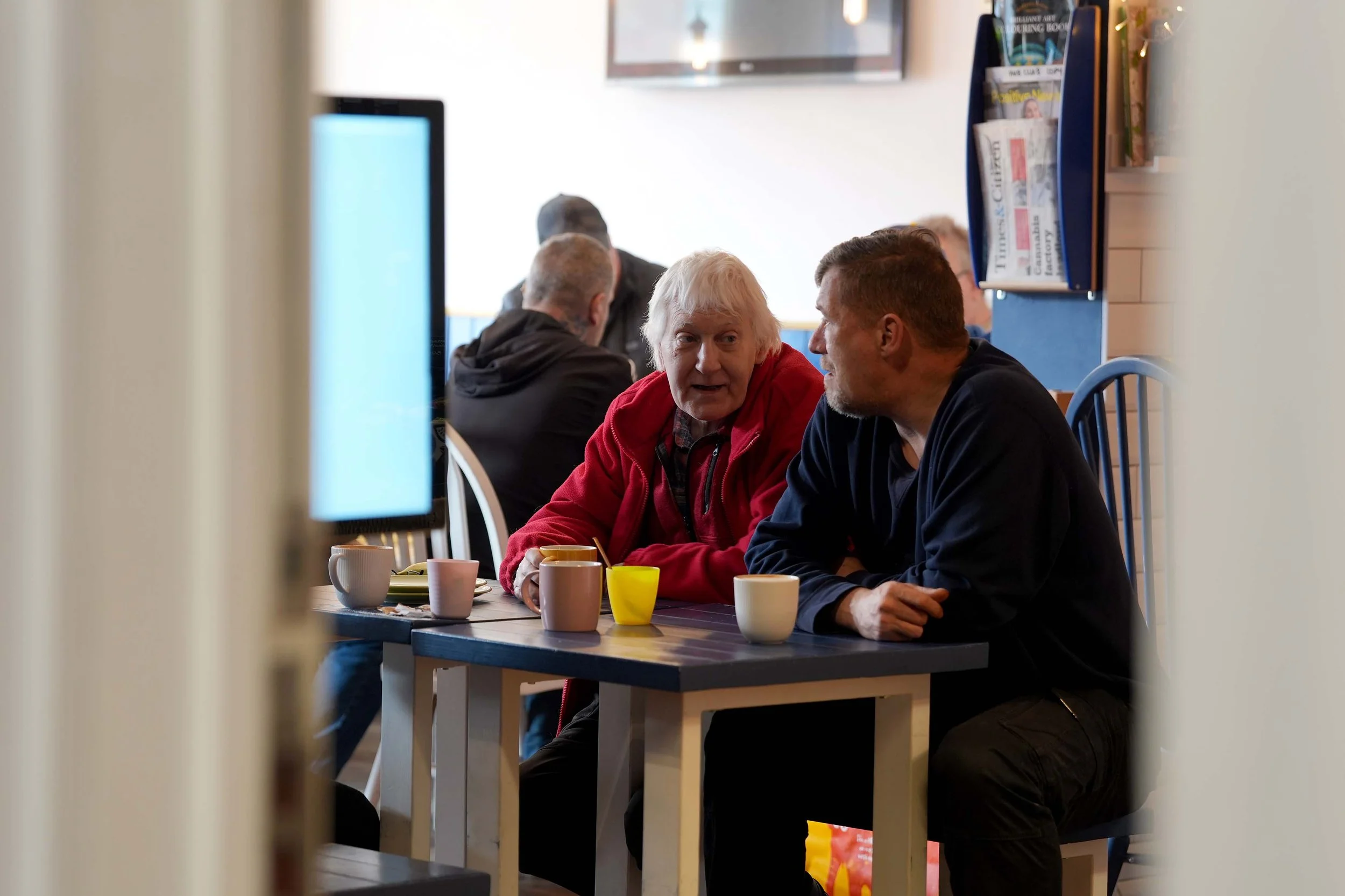 Two men sitting at a table in a cafe, engaged in conversation, with cups and mugs in front of them. The scene is viewed through a doorway or opening, with other customers in the background and takes place at The Shed, a mens group that runs each week