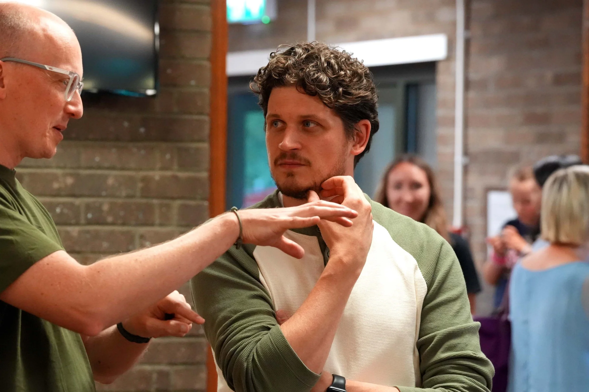 Two young men engage in a conversation, one touching the other's chin gently with a thoughtful expression, in a casual indoor setting with people in the background.